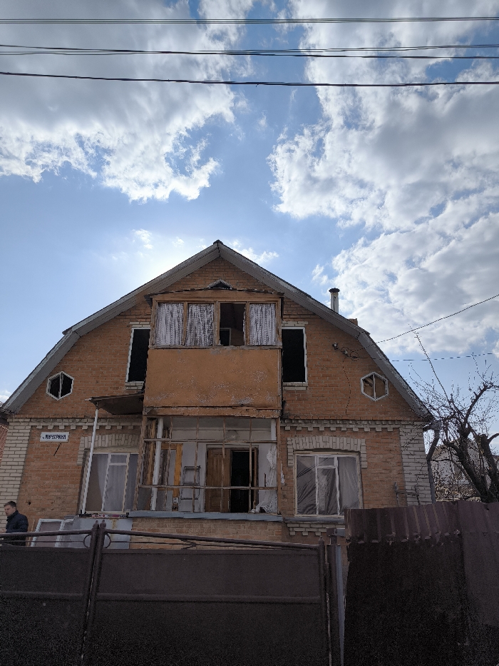 The image depicts the exterior of a two-story brick residential building with an attic, located on Vynnychana Street in Vinnytsia, Ukraine. The structure exhibits significant disrepair. The facade is constructed primarily from red-orange brick, with white brick detailing on the lower portion and corners.
The house features a steeply pitched dark roof with a visible chimney on the right. A central, enclosed balcony on the second story is painted orange-brown, showing extensive peeling paint, and contains three window openings, two of which are covered by tattered white fabric. Below this, an unenclosed lower balcony or porch area reveals exposed wooden framing and open doorways. A wooden ladder is visible leaning inside this lower area.
On either side of the enclosed second-story balcony, large, rectangular window openings are present, appearing empty. In the attic gable, a small, empty rectangular window opening is visible above the central balcony. Additionally, two empty, hexagonal window openings are situated in the attic section, one on each side. Visible cracks traverse the brickwork on the right side of the building. A street sign displaying "ВИННИЧАНА" is affixed to the brickwork on the left side of the house.
In the foreground, a dark, metal gate or fence partially obscures the lower section of the building. To the far left, behind this fence, a single male individual wearing a dark jacket is partially visible. Overhead, multiple utility lines traverse the upper portion of the frame. A bare tree with dark branches is visible on the far right, behind a corrugated metal fence. The sky is partly cloudy with patches of bright blue, and the sun, positioned behind the upper left of the house, casts a bright glare.