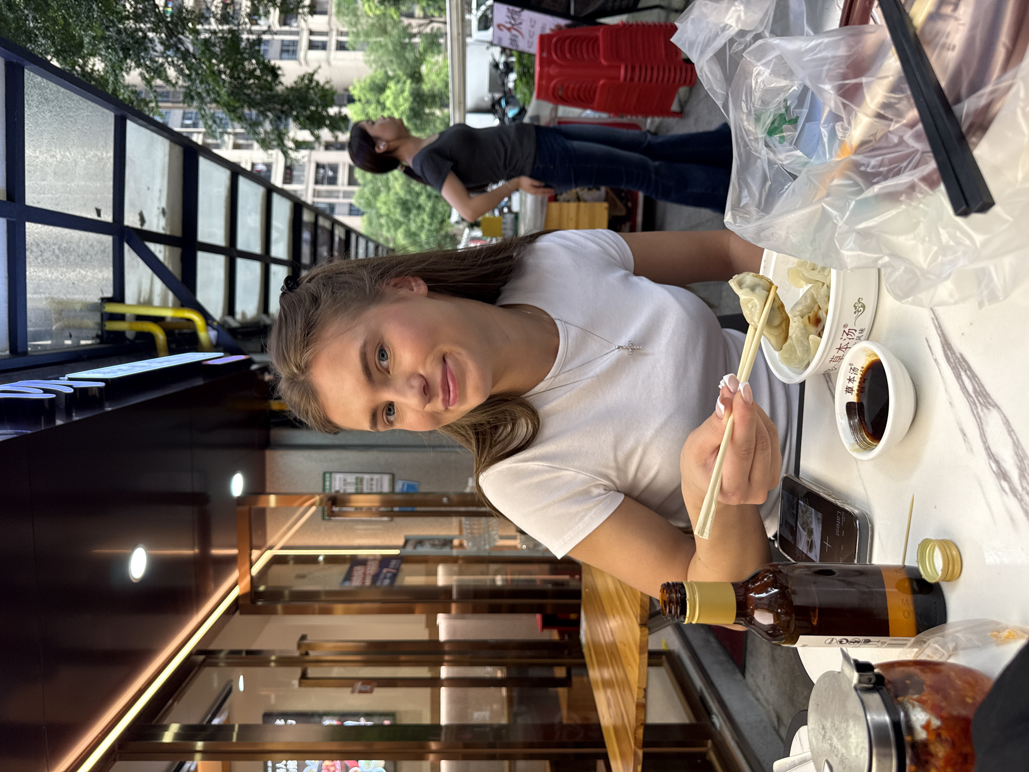 A young Caucasian woman with long brown hair is seated at a wooden table outside a restaurant, holding chopsticks with a dumpling. She is wearing a white t-shirt and a cross necklace. On the table are a bottle of beer, a container of dumplings, a small bowl of soy sauce, a phone, and a jar of chili oil. In the background, another woman with dark hair in a ponytail is walking past, and buildings and trees are visible. The scene appears to be on a street in the Haizhu District of China.