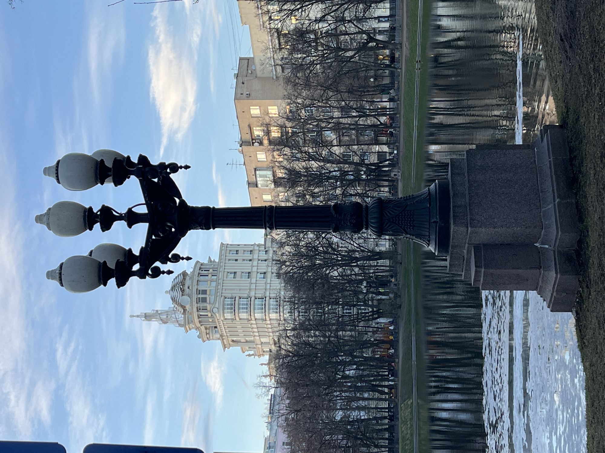 The image shows a street scene in Moscow, Russia, featuring a black ornate lamppost in the foreground. The lamppost has three white, globe-shaped lights. Beyond the lamppost, the view captures a body of water, possibly a river or canal, reflecting the cityscape. Buildings of varying heights and architectural styles are visible in the background, including a tall, white structure with a spire. Bare trees line the waterfront, their branches intricate against the sky. The sky is blue with wispy clouds. The image appears to be taken during the day, with soft lighting illuminating the scene.