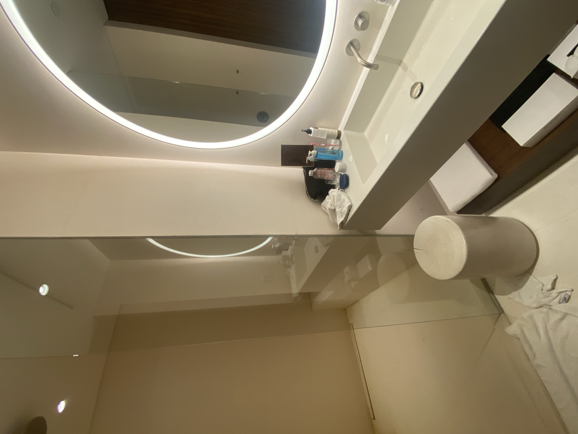 The image depicts a bathroom interior, likely in a hotel or residential setting. There's a brightly lit, round mirror with an illuminated border. Beneath the mirror is a modern, white sink with a stainless steel faucet. A small shelf to the side of the sink holds toiletries.
To the left is a shower enclosed by a glass partition. A white, cylindrical stool sits in front of the glass partition. A white towel is on the floor near the stool. The location of the bathroom is El Caracol, Mexico.