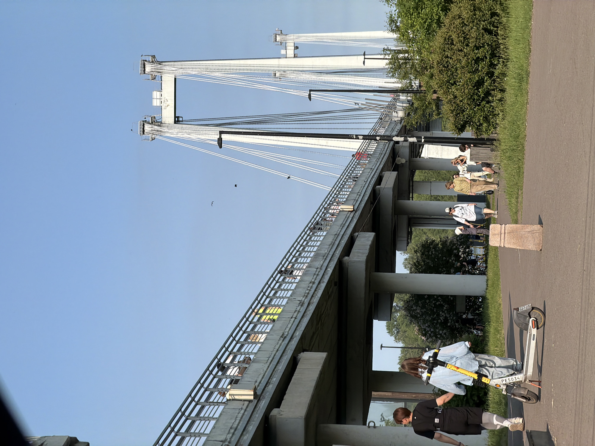 In Krasnoyarsk, Russia, a scene unfolds near a modern cable-stayed bridge under a clear blue sky. Two women walk on a paved path in the foreground, one pulling a white electric scooter. The scooter has yellow and black markings. Behind them, people are visible on the elevated pedestrian walkway of the bridge, which is supported by concrete pillars. The bridge itself is characterized by its white towers and numerous white cables. To the right, dense green bushes and trees form a natural backdrop, bordering the paved area. The overall atmosphere suggests a sunny day in an urban park setting, with the bridge serving as a prominent architectural landmark.