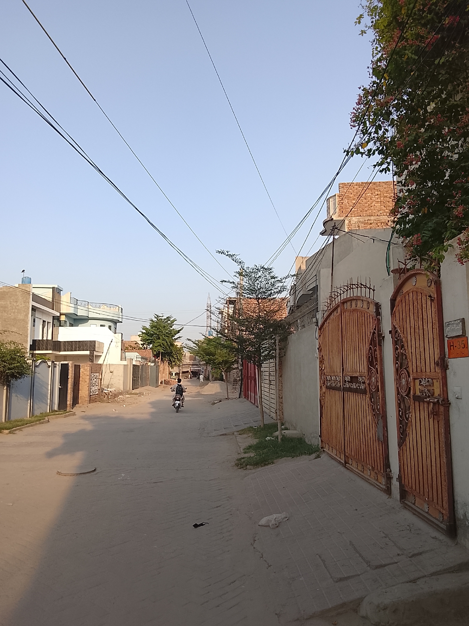 A street in Unknown, Unknown is captured in a full, medium shot during daylight. Overhead, utility wires stretch across the sky. A lone motorcycle with a rider moves down the street. Buildings line both sides of the road, some with brick facades and others painted in light colors. In the foreground, a large metal gate stands to the right. A sign labeled "H-35" is affixed to the wall next to the gate. Debris is scattered on the ground along the street.