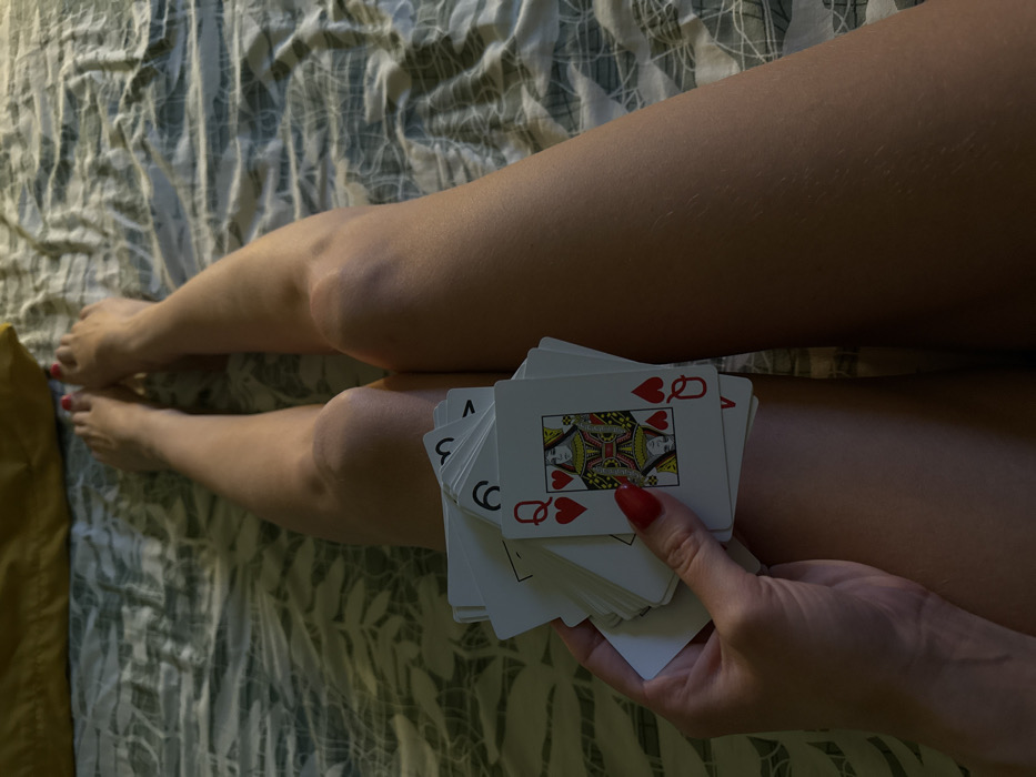 A top-down view depicts a woman's legs resting on a patterned bedspread. A hand, with red nail polish, holds a stack of playing cards, with the Queen of Hearts visible on top. The skin on the legs appears soft and slightly illuminated. The bedspread is patterned in muted greens and whites, creating a relaxed and intimate setting.