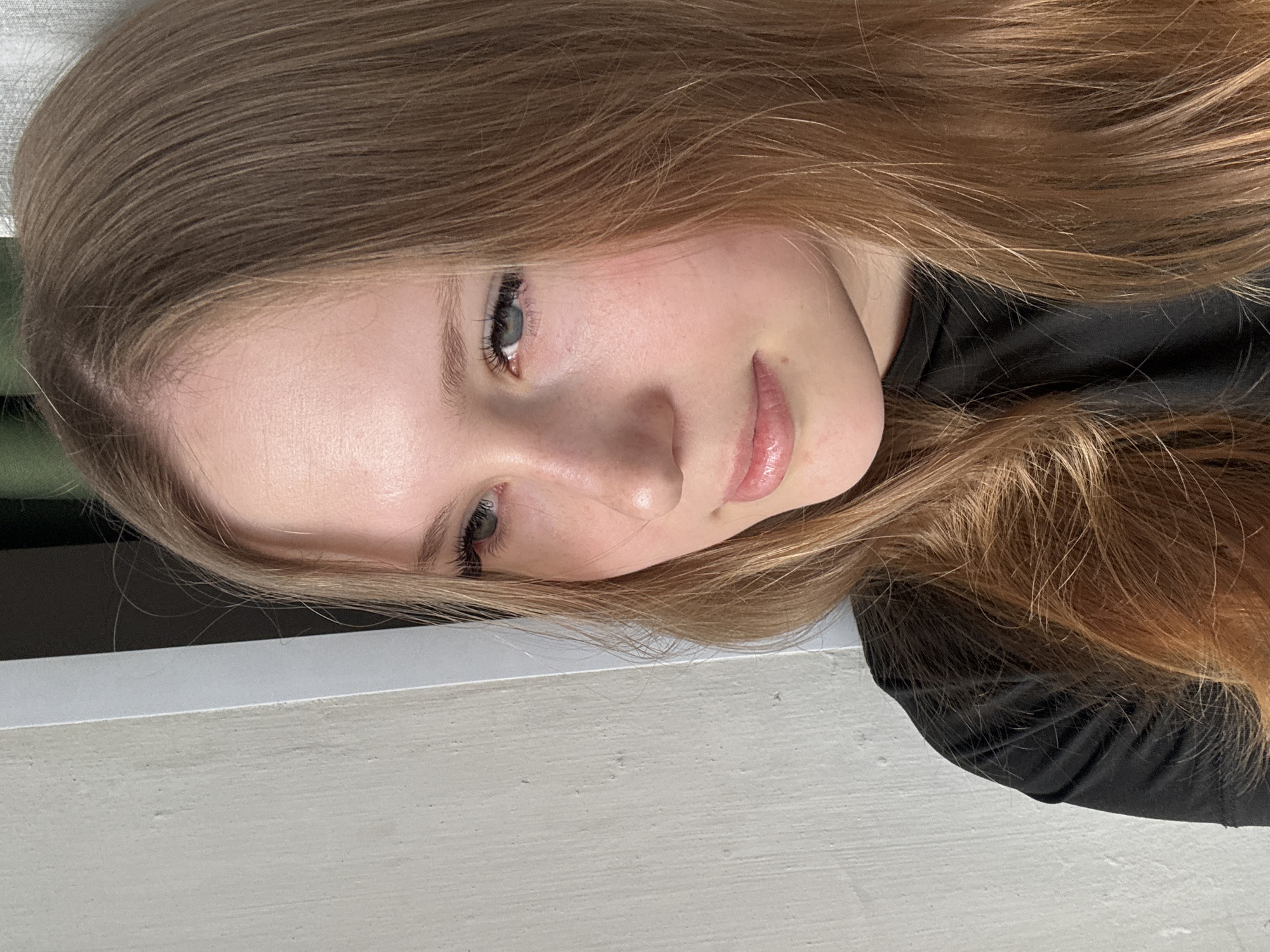 A young woman with light blonde to brown hair, fair skin, and light-colored eyes, poses for a close-up shot. She is wearing a black garment. The background includes a neutral-toned wall and a hint of an indistinct green object. The location, Chaikovsky, Russia, is not discernable from the image content.
