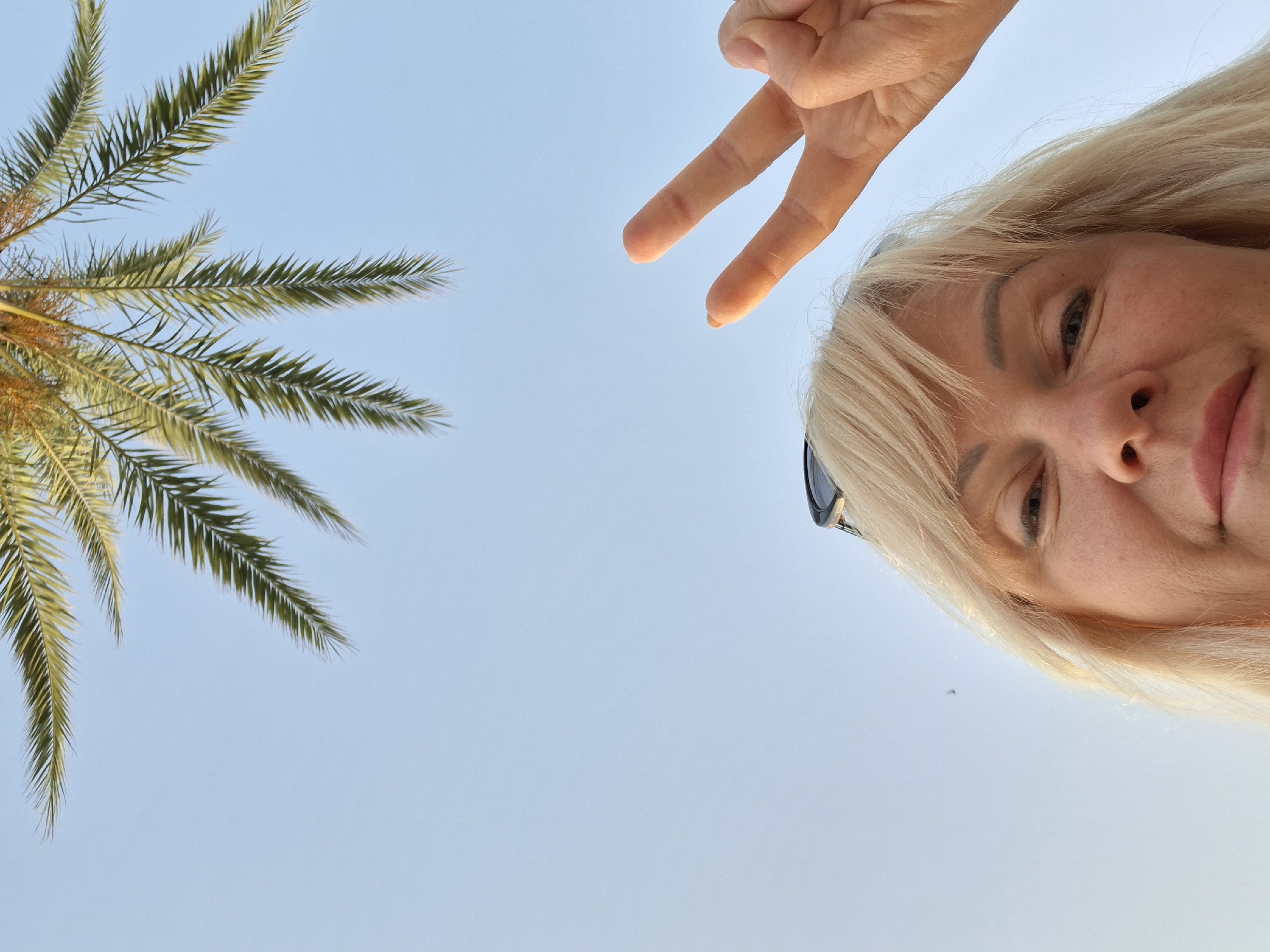 An extreme low-angle, close-up selfie shows a woman with blonde hair and sunglasses on her head looking towards the camera. Her right hand is raised, and she is making a peace sign with two fingers. In the upper portion of the image, palm fronds from a tree are visible against a clear blue sky. The image appears to have been taken outdoors, likely in Torrevieja, Spain, given the presence of palm trees and bright sky.