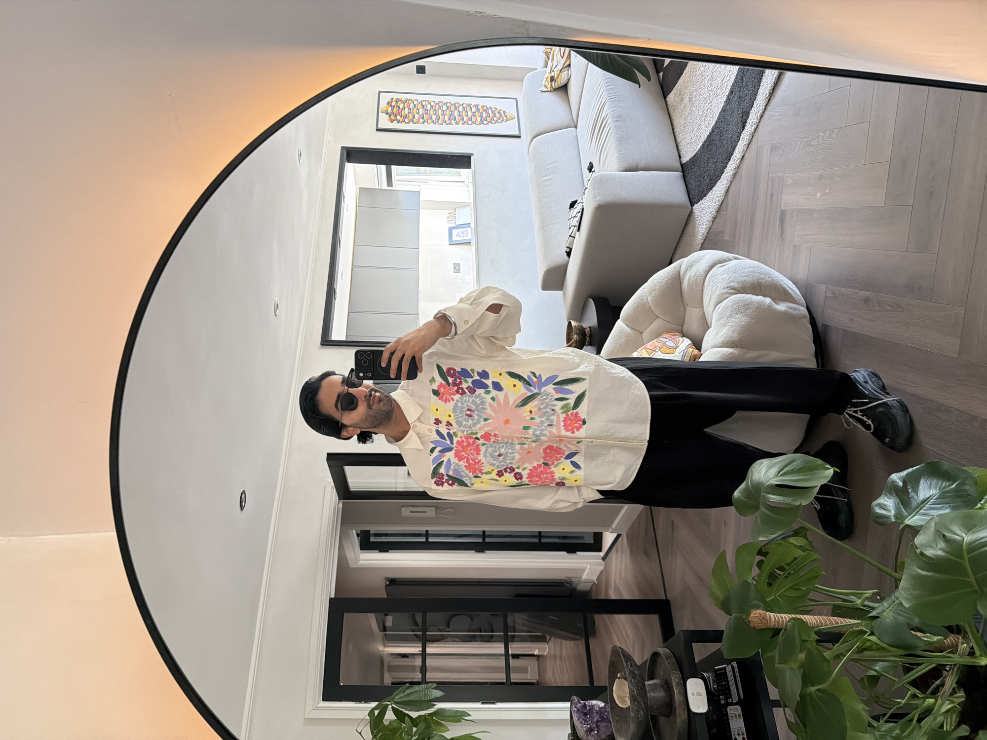 A person is standing in a living room and taking a selfie in a large, rounded mirror. The person is wearing a white long-sleeved shirt with a colorful floral print on the chest and black pants. They are also wearing sunglasses and black sneakers. The living room has a modern design with a light-colored sofa, a unique armchair, and a herringbone-patterned wooden floor. There are also several plants in the room, including a large Monstera plant. A kitchen area is visible in the background through an open doorway. The overall ambiance is bright and airy. The image appears to have been taken in a residential setting.