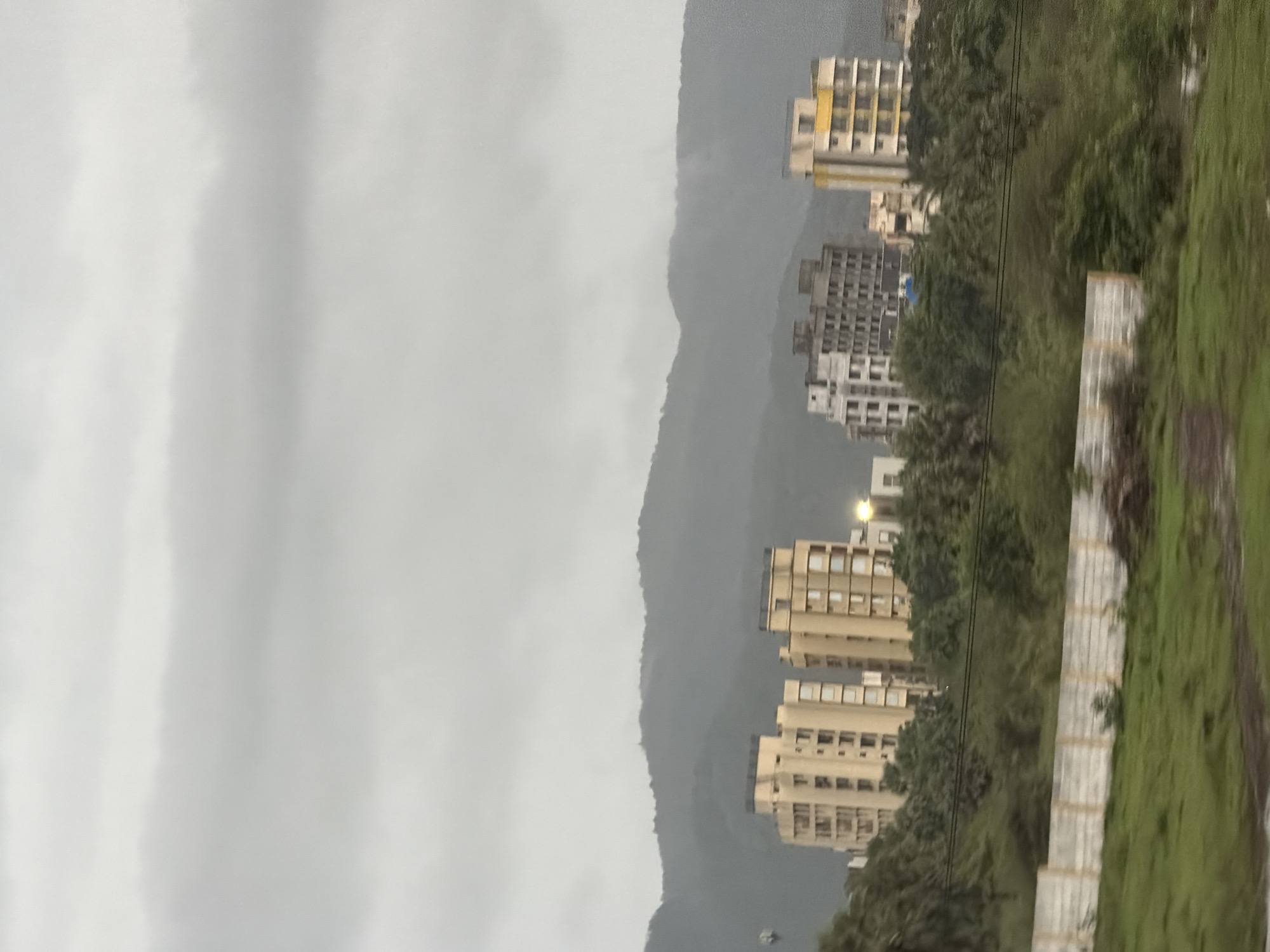 The image captures a cloudy, overcast day in Palghar, India, with a landscape featuring rolling hills in the background. In the midground, several apartment buildings are visible, varying in architectural style and color. The buildings are situated amidst lush green trees and vegetation. In the foreground, there is a grassy area with a low, white concrete wall running horizontally across the scene. The overall impression is one of a developing urban area nestled within a natural, hilly environment.
