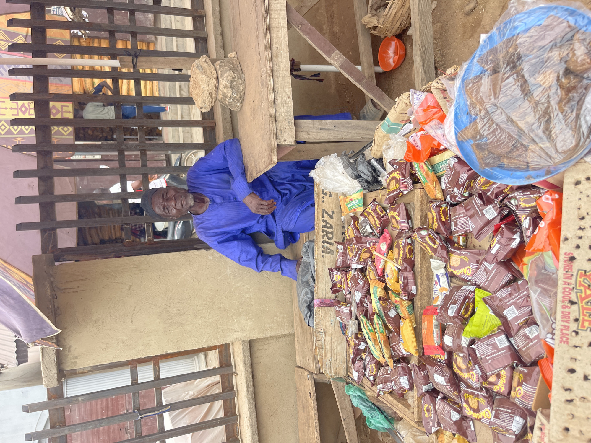 An elderly man in Zaria, Nigeria, is seated behind a table covered with various snacks and baked goods, including "Choco" brand cookies. A blue bowl filled with wrapped food items is also on the table. The table is made of wooden planks and sits in front of a building with a window and metal security bars. Two round rocks sit atop the wooden planking next to the vendor. An orange object is seen on the ground behind the vendor. Text on the product packaging reads "STORE IN A COOL & DRY PLACE" and "NAFDAC REG."