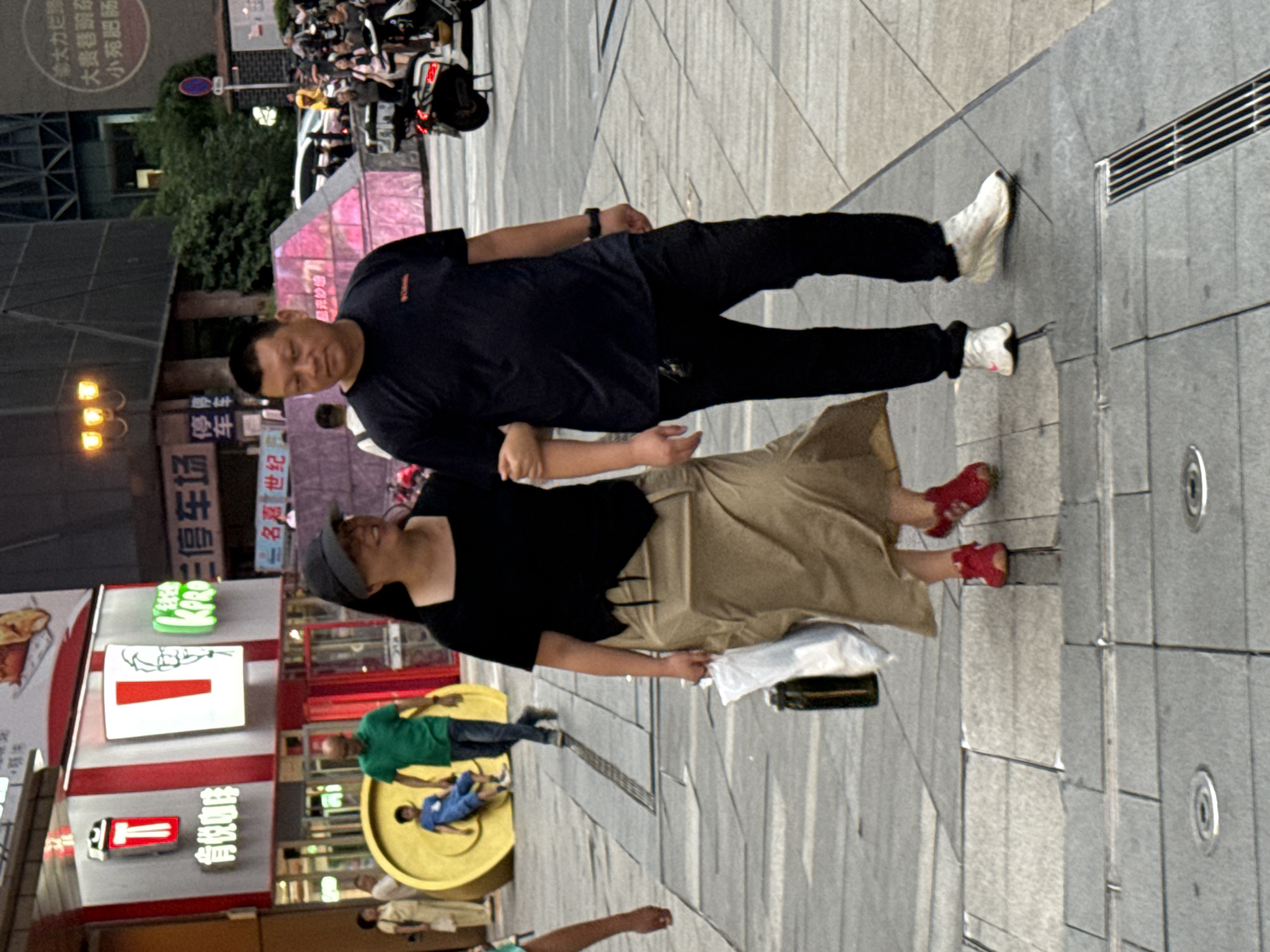 The image captures a street scene in Yuzhong District, China. In the foreground, a man and a woman are walking arm-in-arm. The woman, on the left, wears a black top, a beige skirt, and red shoes. She carries a bag in her left hand and a white shopping bag in her right. The man, on the right, is dressed in a dark blue t-shirt and black pants, and wears white sneakers. They are walking on a paved sidewalk.
Behind them, to the left, is a building with a prominent red and white KFC sign. A man in a green shirt and jeans walks past a large golden circular structure with a child sitting on it. Further in the background, there are other pedestrians, buildings, and some vehicles including a scooter. The overall environment appears to be a busy urban commercial area.