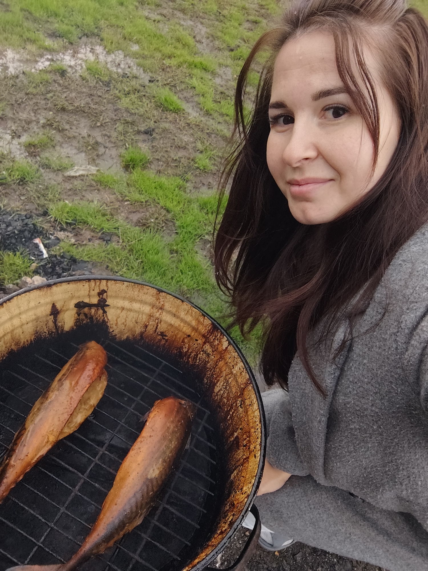 A woman with long brown hair and wearing a gray sweater is smiling and looking at the camera, holding a round grill with two smoked fish on it. The grill is black and coated with dark residue. The background shows a grassy area, with some patches of mud, suggesting a damp or recently wet environment. Based on the information provided, there is no information available to confirm this image was taken in the city of Фоки, Russia.