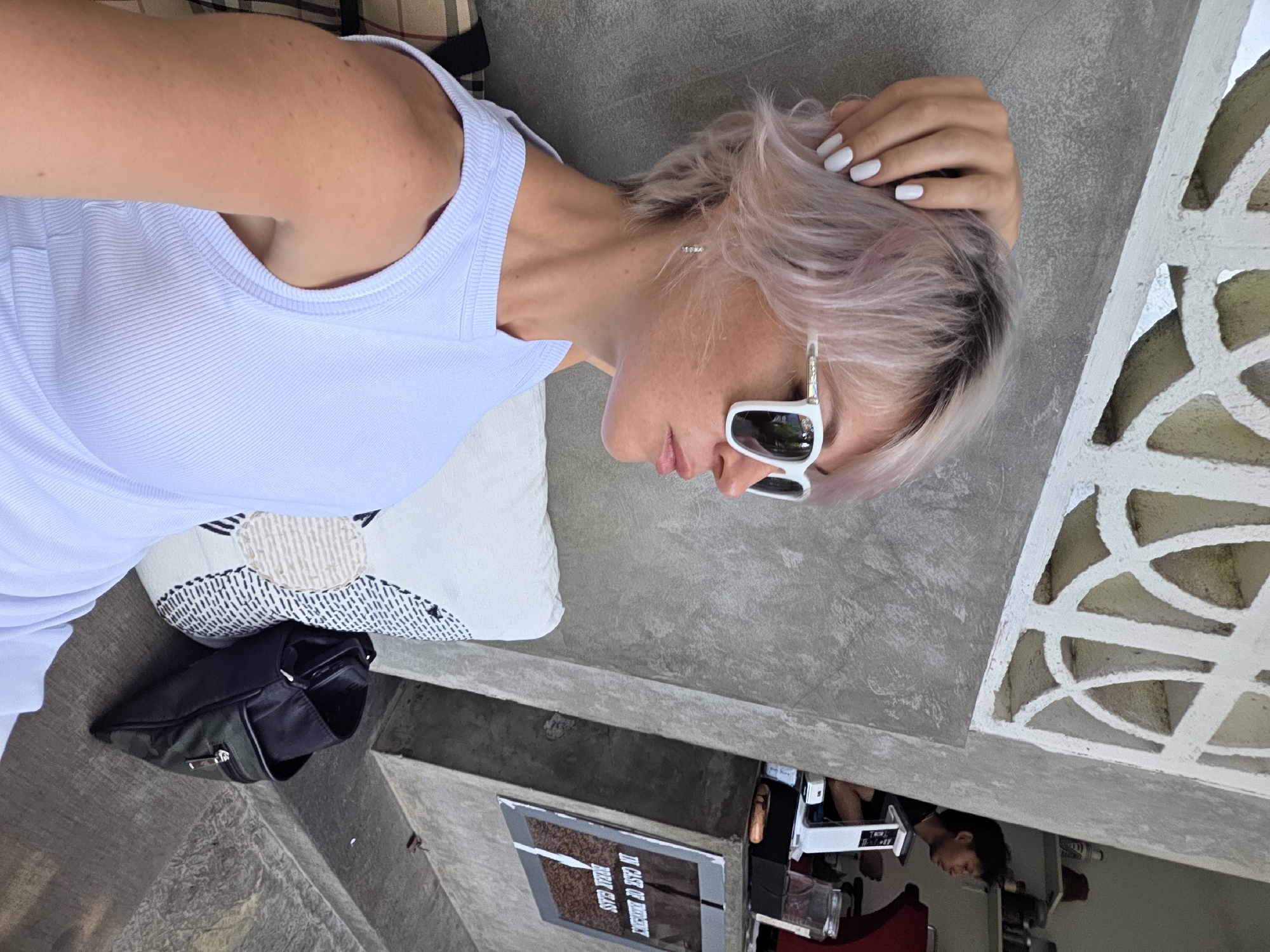 An outdoor, eye-level, medium shot captures a woman with short, blonde-pink hair wearing a white tank top and white sunglasses. Her right hand rests on her head. She is seated next to a patterned cushion. Behind her, a concrete wall features an abstract cutout design. In the background, a person with dark hair works at a coffee counter, with shelves of items visible. A framed sign with text that reads "IN CASE OF EMERGENCY BREAK GLASS" is mounted on a slanted surface. A dark green bag is placed beside the cushion. The scene appears to be set in a cafe in North Kuta, Indonesia.