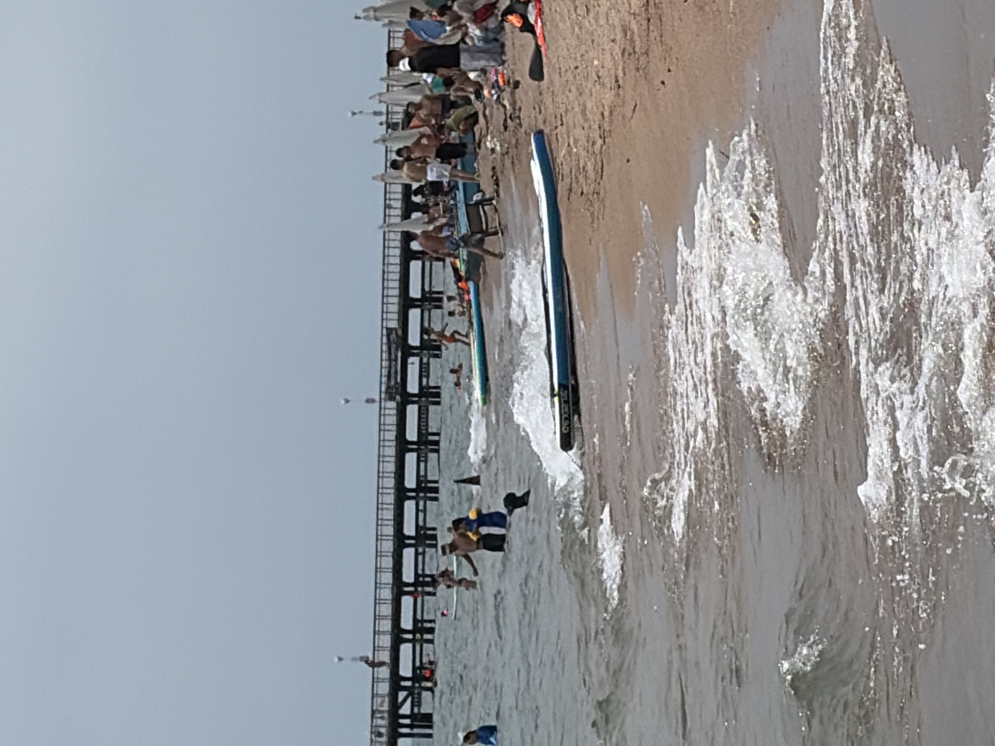 The image depicts a crowded beach scene with people in and around the water. A long pier extends into the ocean in the background, with a cloudy sky above. Several individuals are swimming or wading in the water, while others are standing or sitting on the sandy shore. Two stand-up paddleboards are visible, one in the water and another on the beach. Beach umbrellas are set up on the sand, and some people are gathered around them. The waves are gently breaking on the shore. The overall atmosphere suggests a busy day at a beach resort.