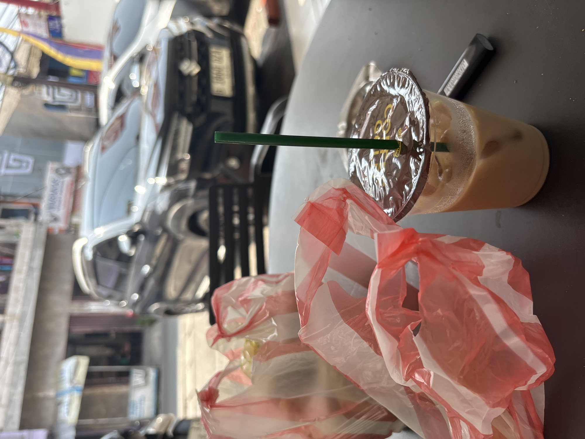 A close-up, eye-level shot depicts a dark-colored table with various objects on it, set against an urban background. In the foreground, to the left, are two red and white striped plastic shopping bags, one slightly behind the other, containing some items. To the right, a clear plastic cup with a dark-colored lid holds a light brown beverage with ice and a green straw. Next to the cup, a black object, possibly a marker or pen, rests on the table. In the blurred background, a dark grey SUV is parked on a street, with buildings and other vehicles visible. The scene appears to be outdoors, possibly in a commercial area of North Jakarta, Indonesia, suggested by the Indonesian license plate on the SUV and the general streetscape. No people are visible in the frame.