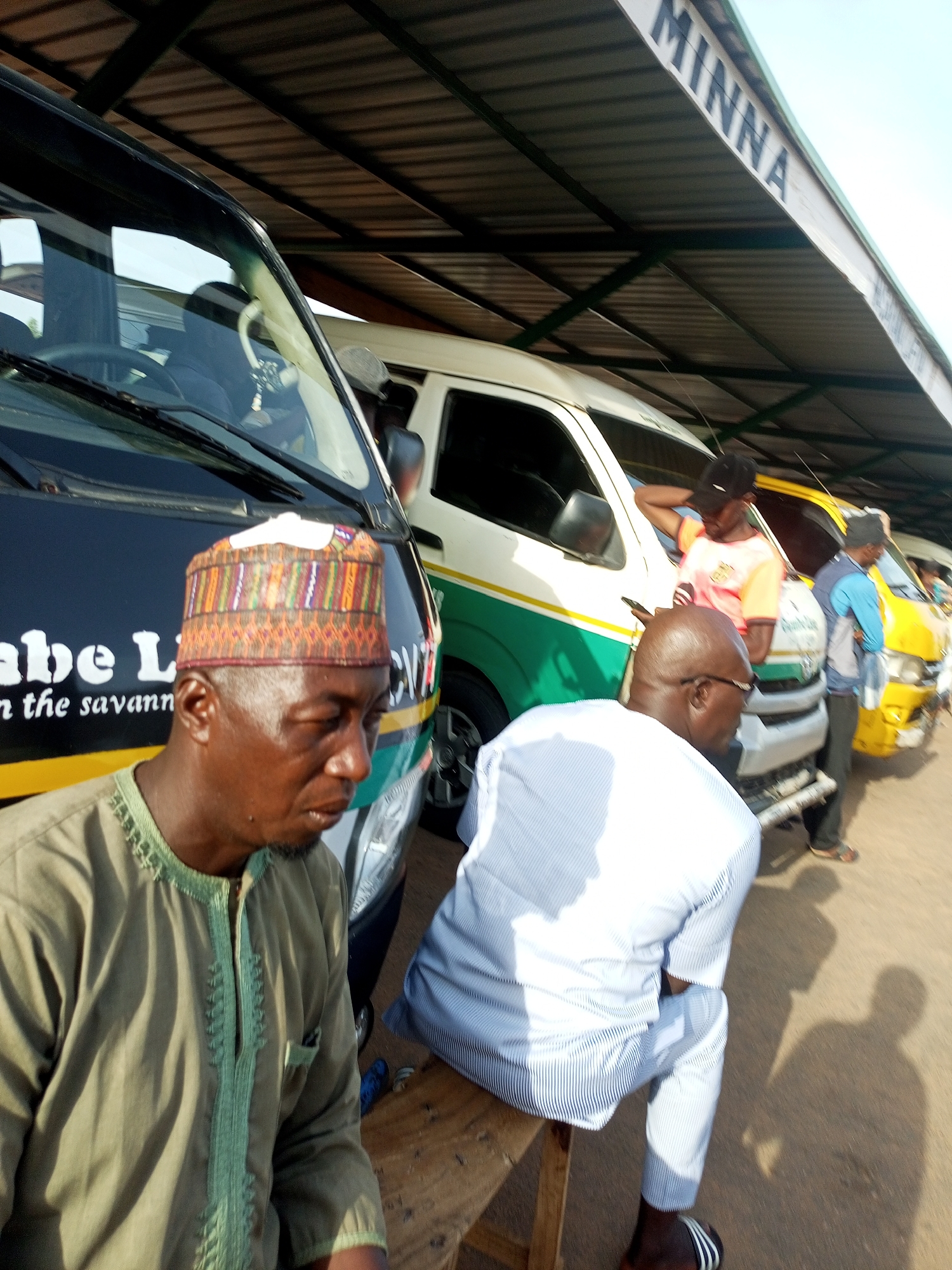 The image depicts an outdoor scene under a covered walkway, possibly a bus station. A man wearing a colorful, patterned cap and a green, embroidered tunic is seated in the foreground, facing right. He is looking down and to his right. Behind him, other individuals are visible near several vehicles. To the left, a black and white bus is parked, with lettering on its side. To the right, a white and green bus is parked, followed by a yellow bus. The walkway has a corrugated metal roof supported by dark metal beams. The word "MINNA" is visible in black capital letters on the side of the walkway's roof. The ground appears to be dirt or sand.