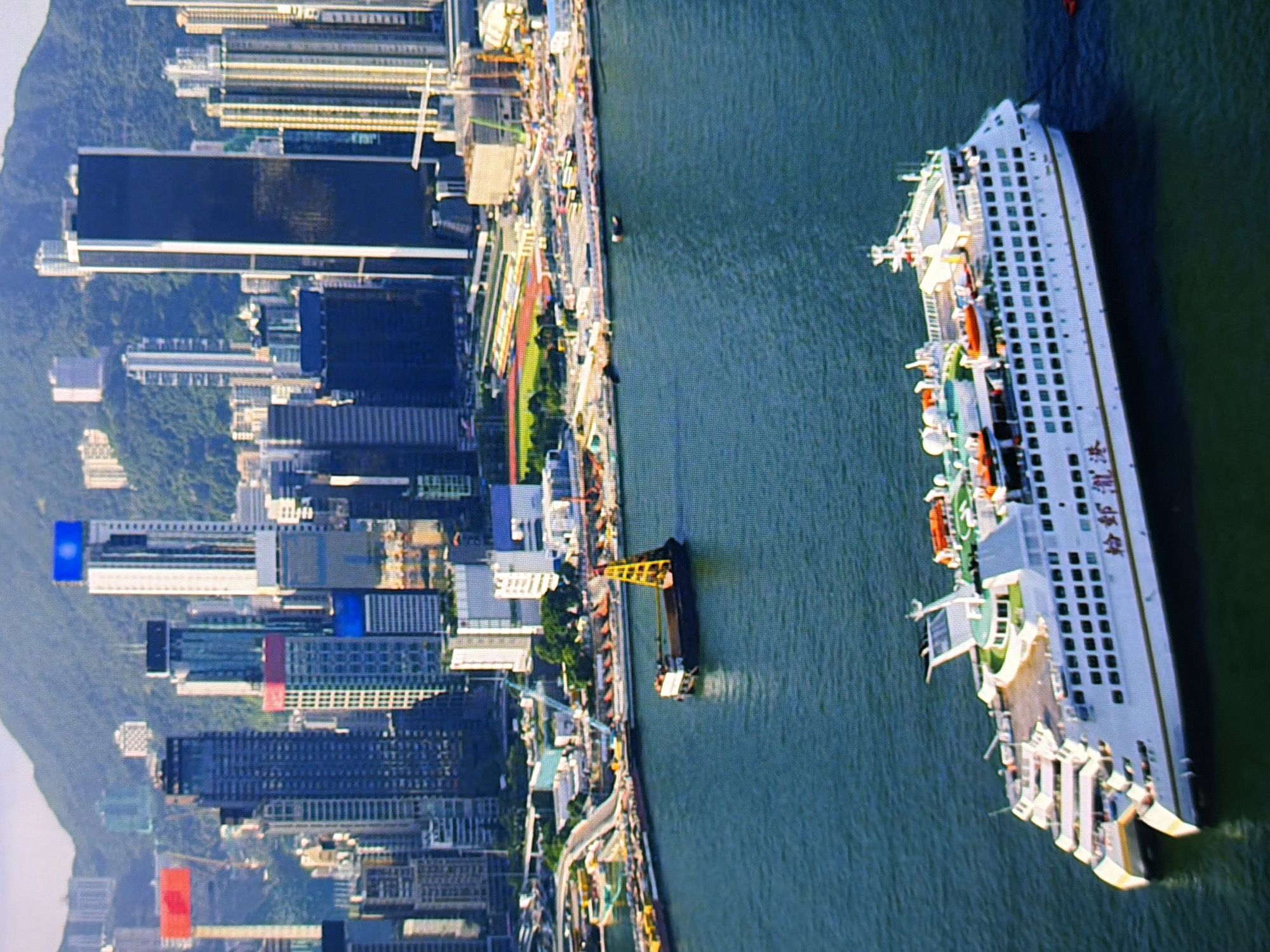The image shows a wide shot of a harbor with a modern ferry docked in the foreground. The ferry is white with several decks and has Chinese characters written on its side. In the background, a dense urban landscape with numerous skyscrapers rises against a mountainous backdrop. The sky is partly cloudy, and the water is a deep blue-green. Construction cranes are visible along the waterfront, suggesting ongoing development. The scene is set in a bustling port city, but it is not Kyiv, Ukraine, as Kyiv is an inland city and does not have a harbor of this nature.
