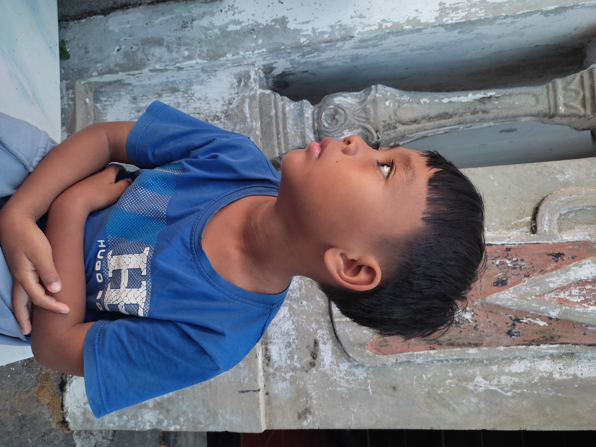 A young boy with a bowl cut and dark hair is sitting outdoors, looking to his left. He is wearing a blue t-shirt with a white design on it, and light gray pants. His arms are crossed in front of him. The boy is positioned in front of a weathered concrete wall with some faded orange and white markings, and a decorative concrete pillar to his left. The scene suggests an outdoor setting in a residential or public area. The visual details are consistent with an environment found in Ponorogo, Indonesia, given the architectural style and the boy's appearance.