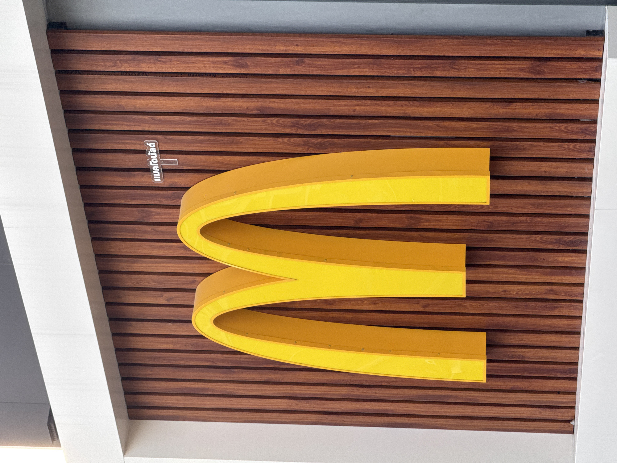 A close-up, eye-level, exterior shot captures a large, three-dimensional golden "M" logo, characteristic of McDonald's, mounted on a vertical wooden plank wall. The planks are dark brown with visible grain. Above and to the right of the "M," a small, clear sign with Thai lettering is affixed to the wall. The overall scene is bright, indicating daylight. The location is identified as Ban Tai Subdistrict, Thailand.