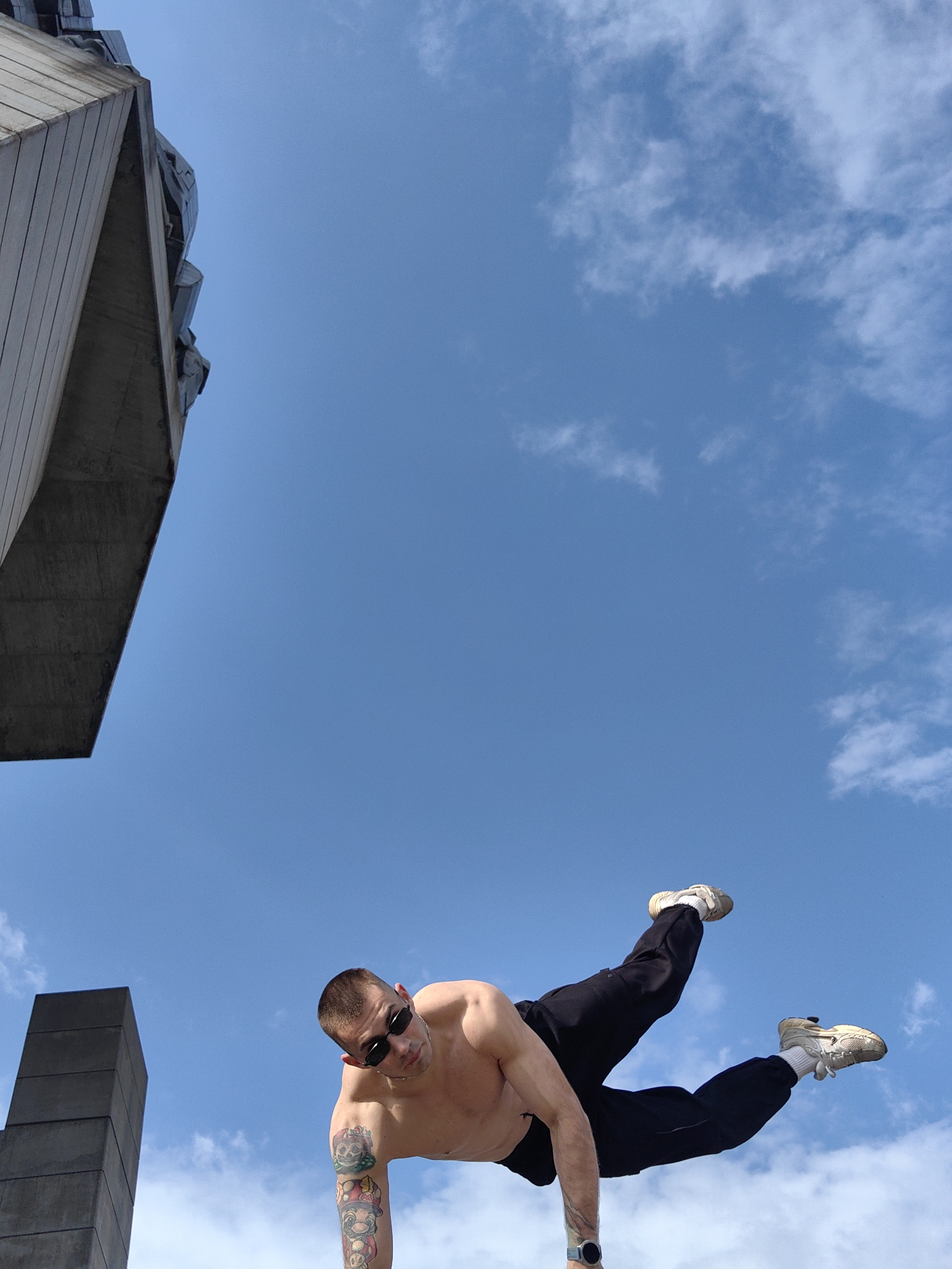 A low-angle, medium shot captures a shirtless man with short hair and sunglasses, performing a handstand in front of a clear blue sky with scattered clouds. He wears black track pants and white sneakers. A large concrete structure occupies the upper left corner, while a stacked concrete pillar is visible in the lower left. This location is in Shumen, Bulgaria. The man has tattoos on his left arm and wears a watch on his left wrist.