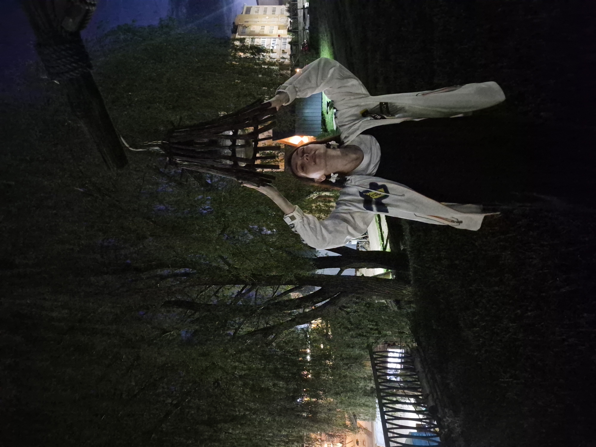 A young person stands in front of a wooden structure with illuminated interior at night in Votkinsk, Russia. The person is wearing a black shirt with a white jacket. The background includes a residential building and greenery. A metal fence is partially visible.