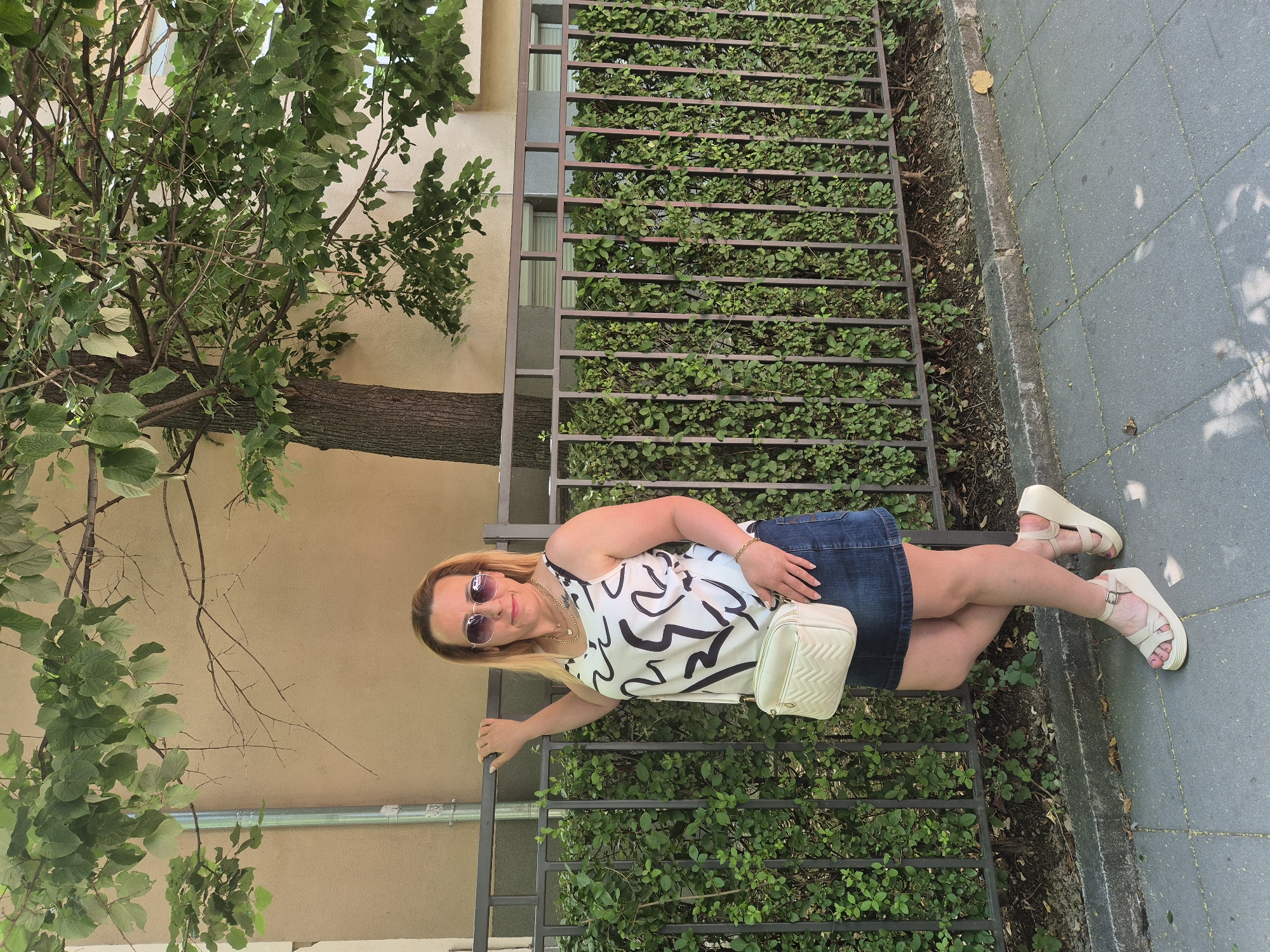 A blonde woman with sunglasses and a white patterned tank top stands next to a metal fence covered in green bushes. She is wearing a denim skirt, a white quilted crossbody bag, and beige wedge sandals. Her left hand rests on the fence, and her right arm is bent at the elbow, with her hand near her hip. Behind the woman, a building with a tan facade and some trees with green leaves are visible. The scene appears to be on a sidewalk with a paved surface and a stone curb.