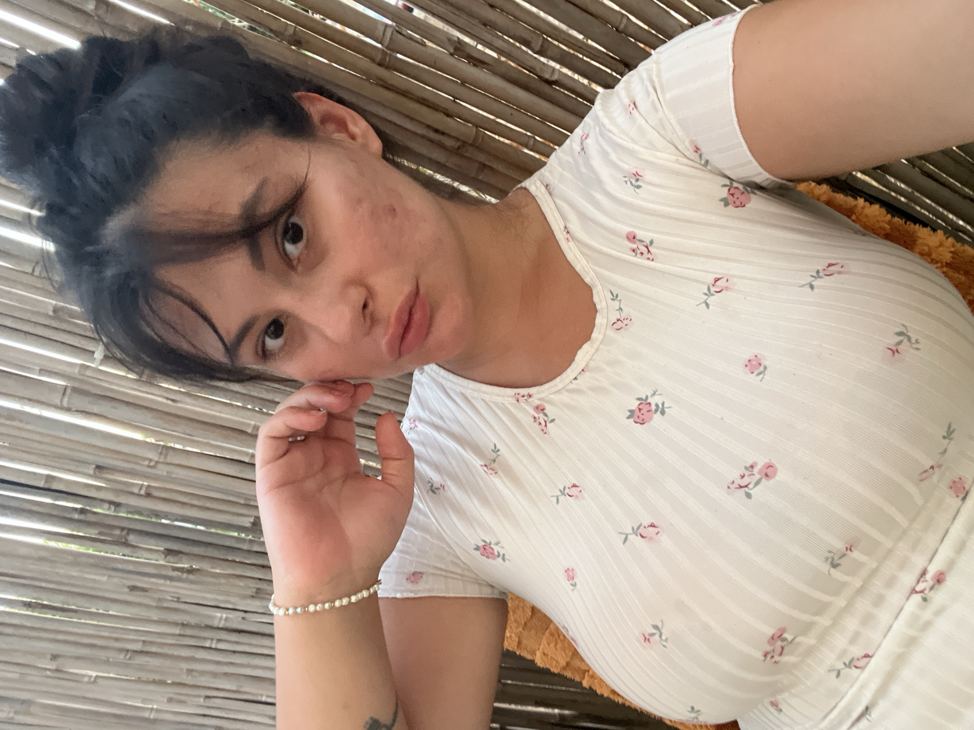 A young woman with dark hair pulled up in a bun and bangs framing her face is looking at the camera with her hand near her cheek. She is wearing a white, short-sleeved shirt with a subtle floral pattern of pink and green flowers. She has a pearl bracelet on her left wrist. The background appears to be a wall made of vertical bamboo poles. The setting suggests an outdoor or semi-outdoor environment. No specific location in Kiryat Yam, Israel, can be identified from the image alone.