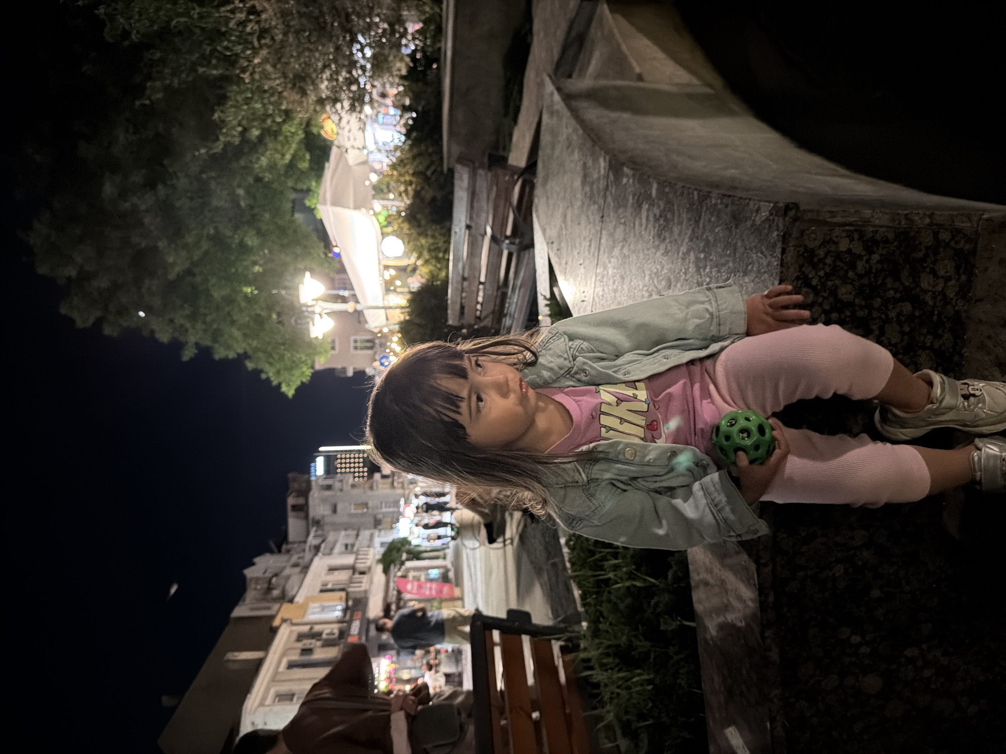 A young girl sits on a stone bench outdoors at night, holding a green ball. She is wearing a pink t-shirt, a light blue denim jacket, pink leggings, and silver sneakers. The background shows a street scene in Burgas, Bulgaria, with buildings, streetlights, trees, and a closed outdoor seating area of a restaurant. There are other people in the background, but they are out of focus. The overall scene suggests a relaxed evening in a public space.