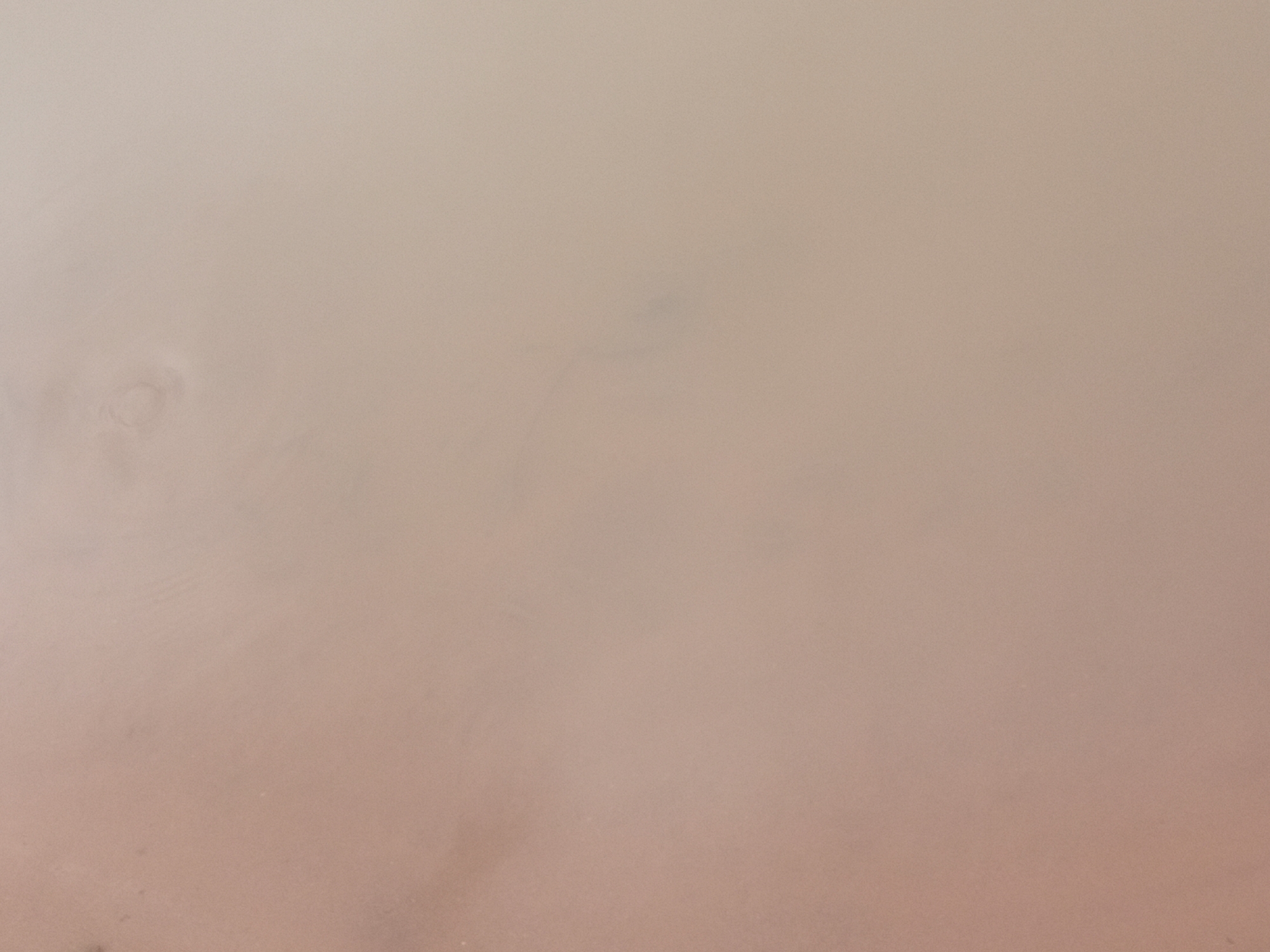 The image depicts a close-up, aerial view of a sandy surface with subtle textural variations and color gradients. The dominant colors are light brown and beige, suggesting dry earth or sand. There are faint circular patterns and slight undulations in the surface, possibly indicative of wind patterns or the drying of mud. In the upper left quadrant, a slightly darker, more defined circular impression is visible, resembling a shallow imprint or disturbance in the sand. No people, objects, or actions are discernible in the image. The scene is devoid of any context that would suggest a specific urban environment, including Nanka, Nigeria.
