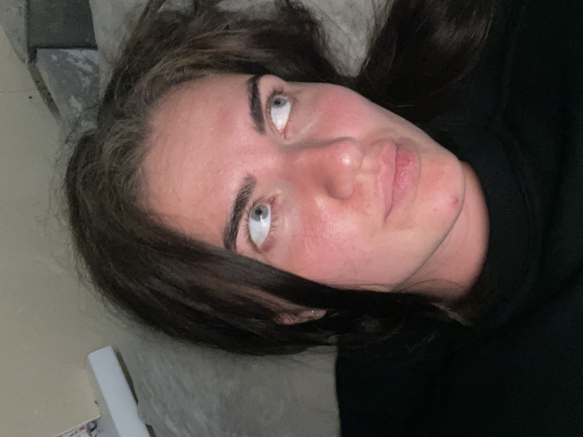 The image shows a close-up, overhead shot of a young woman's face. She has dark, wavy hair framing her face, and her skin appears flushed. Her eyes are a light blue-green color, and she is looking off to her right. Her lips are slightly parted. She is wearing a dark-colored garment, possibly a shirt or sweater. The background is out of focus and appears to be a textured surface, possibly a wall or bedding. The lighting is dim, casting shadows on her face. There is no discernible information about the location in Мосметаллоконструкция Онт, Russia.