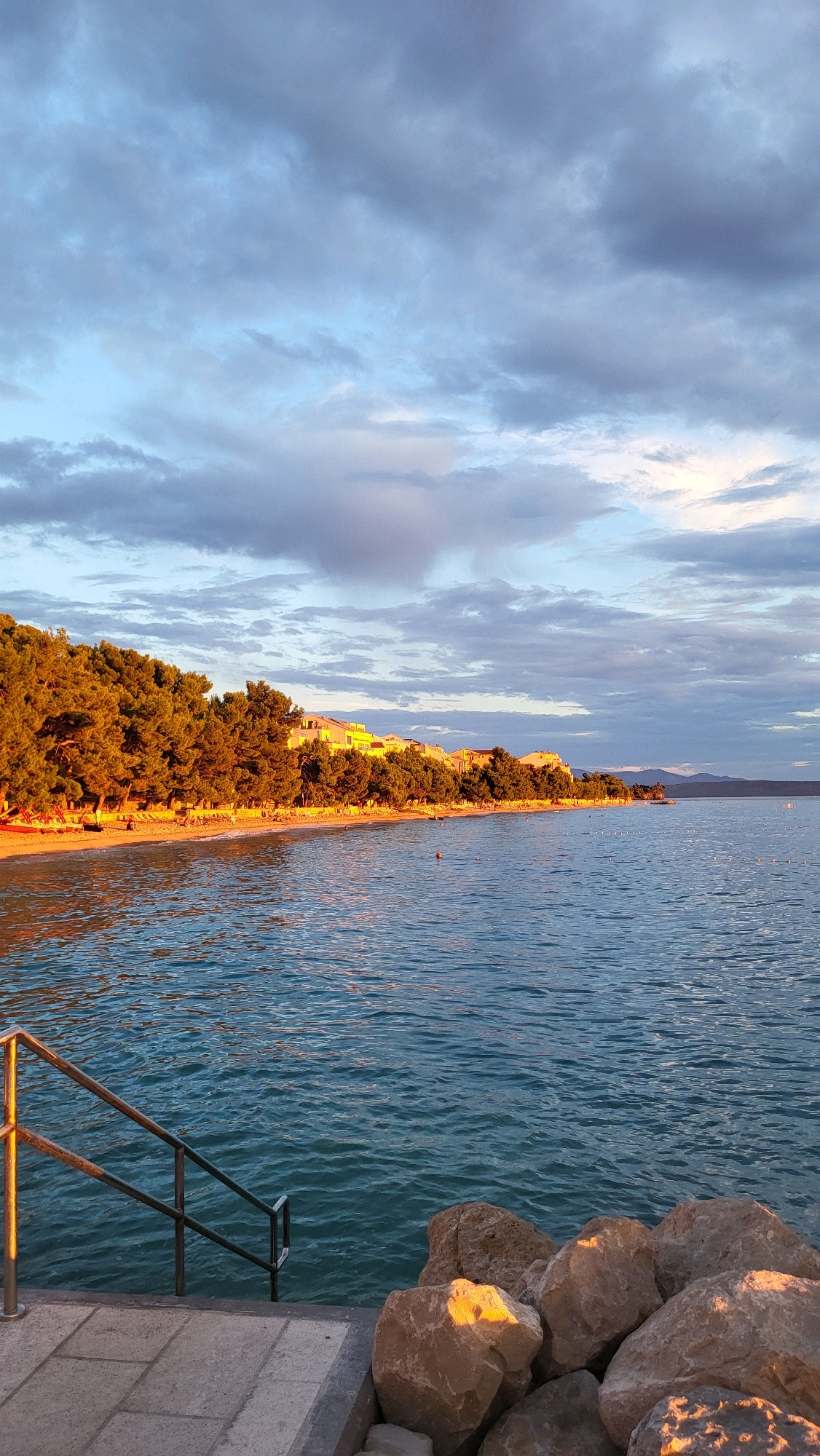 A vertical, eye-level shot captures a tranquil beach scene in Općina Tučepi, Croatia, during what appears to be the golden hour. The foreground features a paved walkway with square tiles, leading to a set of metal railings that descend into the water. Adjacent to the walkway, a cluster of large, light-brown rocks is partially illuminated by the setting sun. The calm, turquoise water of the sea fills the mid-ground, reflecting the warm glow of the sky. In the distance, a crescent-shaped sandy beach is lined with a dense growth of tall, green pine trees, their foliage bathed in the warm, orange light. Behind the trees, a row of light-colored buildings, characteristic of a coastal town, stretches along the shoreline. The sky above is a blend of soft blue and gray clouds, with hints of pink and orange from the sunset. The overall atmosphere is serene and picturesque, typical of a Mediterranean coastal resort.