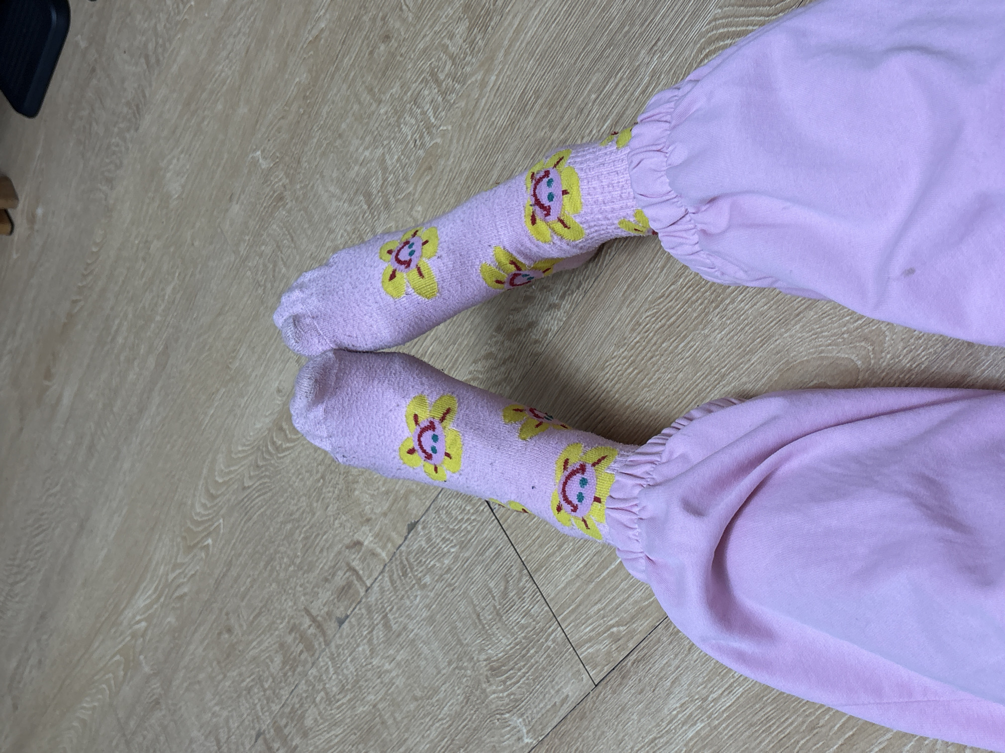 The image shows a pair of legs and feet on a light wood-grain floor. The legs are covered in pink clothing that bunches at the ankles. The feet are covered in light purple socks with a repeating pattern of yellow flowers that have faces. The location is not discernible from the image content.