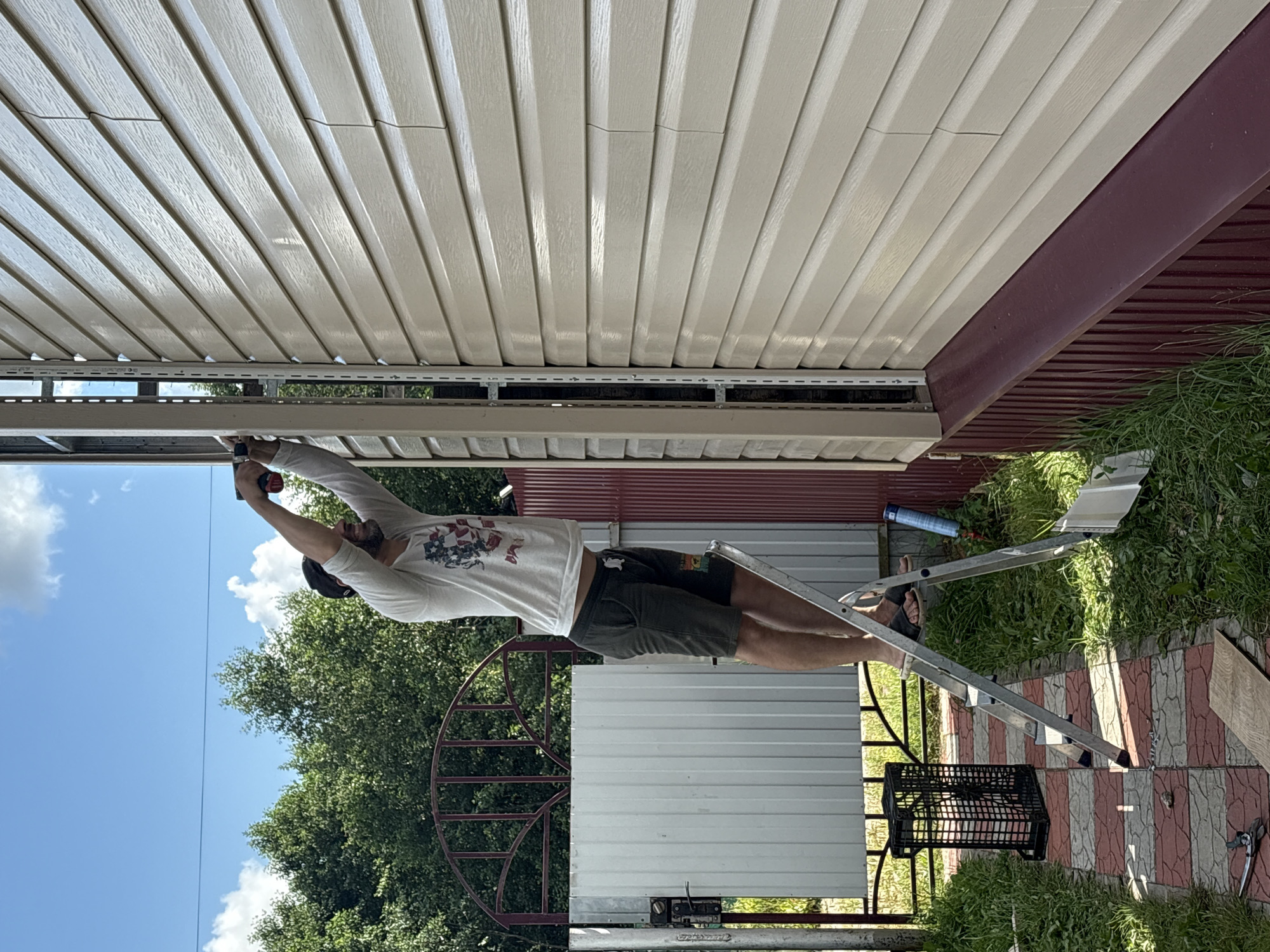 A man stands on a step ladder, performing exterior home repairs. He is wearing a white t-shirt with a graphic design and dark shorts. His right arm is raised, and he is using a power drill to attach a piece of siding to the side of a beige-colored house. The house has a dark red trim along the bottom. To the left of the house, a metal gate and fence are visible, along with a black plastic crate. The ground is covered with green grass and a red and white checkered pathway. Trees and a blue sky with scattered clouds are in the background. The scene appears to be outdoors, in a residential area, likely in Ветлужский, Russia, given the context provided.