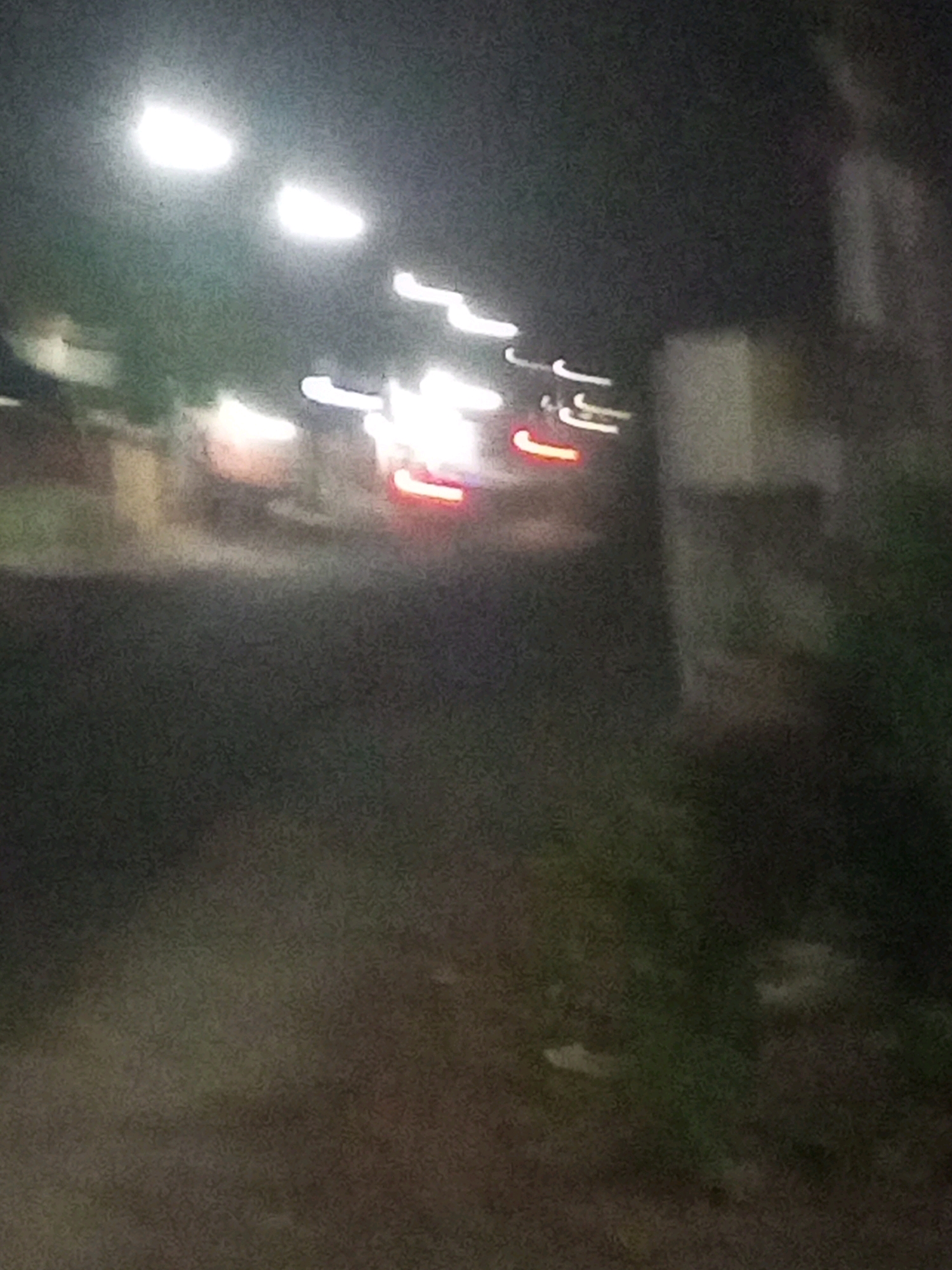 The image depicts a blurry, nighttime scene on a street in Ngasem, Indonesia. The focus is on a moving vehicle, likely a car, with its headlights and taillights blurred due to motion and low light conditions. The street appears to be paved and there are some indistinct structures on either side, possibly buildings or trees. The overall impression is one of a transient moment captured at night.