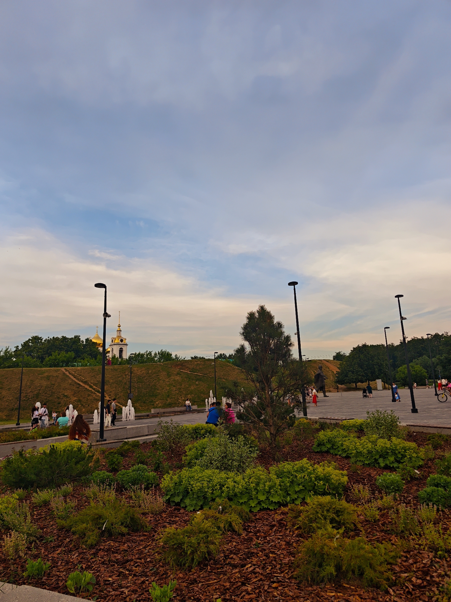 The image depicts an outdoor park scene with a variety of elements. In the foreground, there is a landscaped garden bed filled with mulch and various green plants, including bushes and ground cover. Beyond the garden, a paved area is visible, with several people walking and standing around. Some are engaged in conversation, while others are observing their surroundings.
A prominent feature in the mid-ground is a series of fountains, with water spraying upwards. Behind the fountains, a grassy slope rises, topped with a building that has golden domes, suggesting a church or religious structure. Trees and vegetation line the top of the slope.
Further back, more trees form a backdrop, and in the distance, a cloudy sky with hints of sunset colors stretches across the frame. Several lampposts are strategically placed throughout the park, illuminating the pathways and open spaces. On the right side of the paved area, a person is seen riding a bicycle. The overall impression is that of a public park in a city, likely during the late afternoon or early evening, with people enjoying recreational activities and the ambiance of the park.