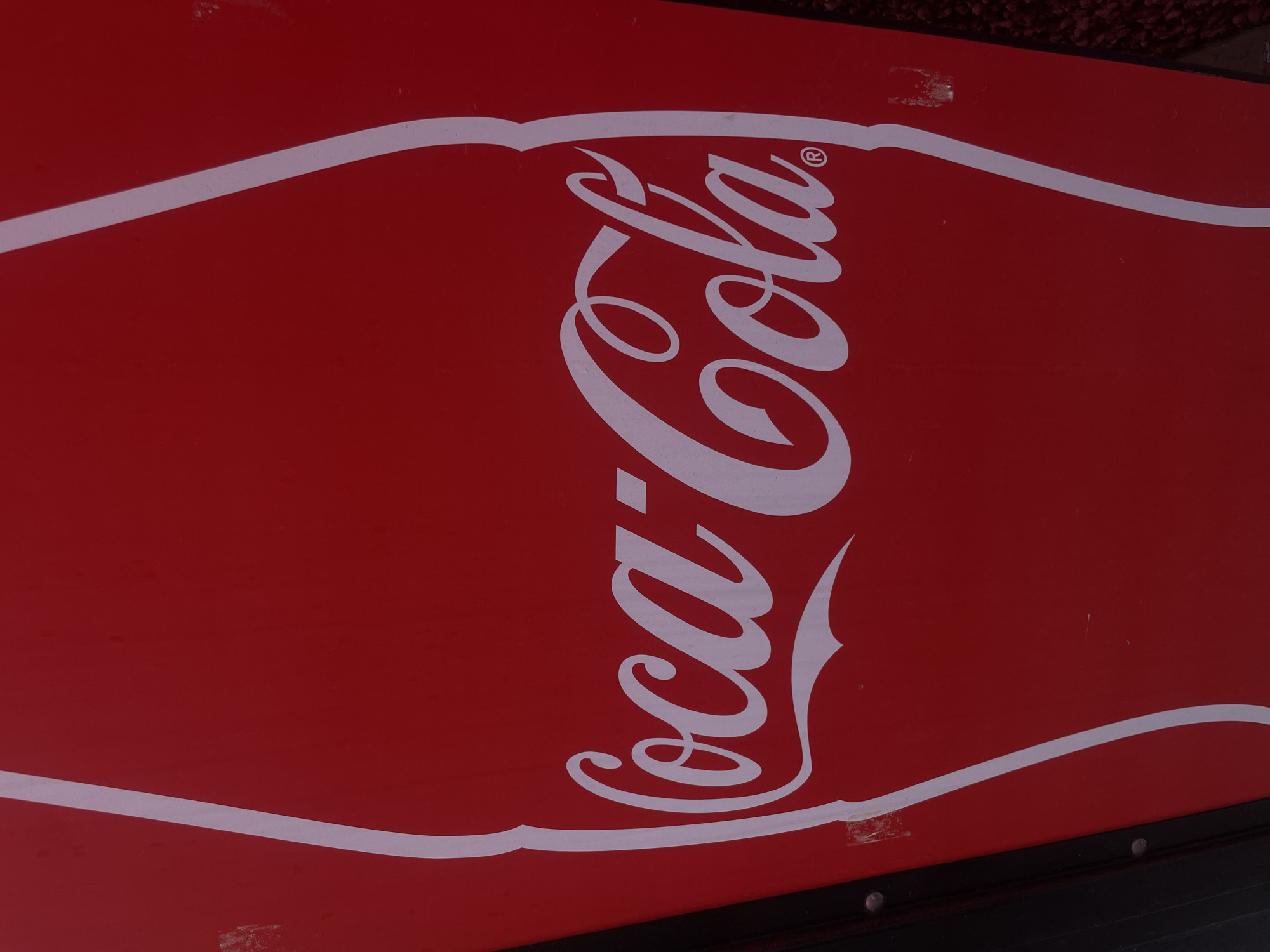 The image displays a close-up, vertical shot of a vibrant red sign featuring the iconic white "Coca-Cola" script logo. White outlines, suggesting the contour of a Coca-Cola bottle, are partially visible along the left and right edges of the red surface. The "Coca-Cola" lettering, including a registered trademark symbol (®) adjacent to "Cola," is centrally positioned within the frame. The sign exhibits minor surface wear, including some scuff marks, particularly on the left side near a dark, possibly black, border secured with visible rivets. To the immediate right of the red sign, a blurred, textured surface, possibly reddish-brown, is discernible. The scene is located in San Andrés Cholula, Mexico. No individuals are present in the image.