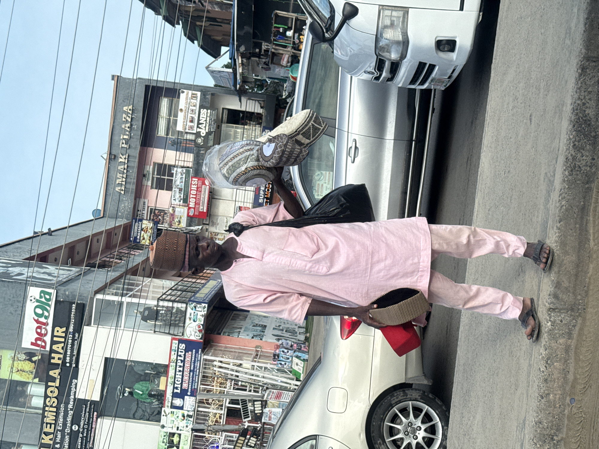 A man stands on the sidewalk next to parked cars in Ado, Nigeria, selling traditional Hausa caps. He is carrying a bag on his shoulder and holding caps. Behind him are buildings with storefronts, with signs for businesses like Kemisola Hair, Lightplus Electrical, and Amak Plaza.