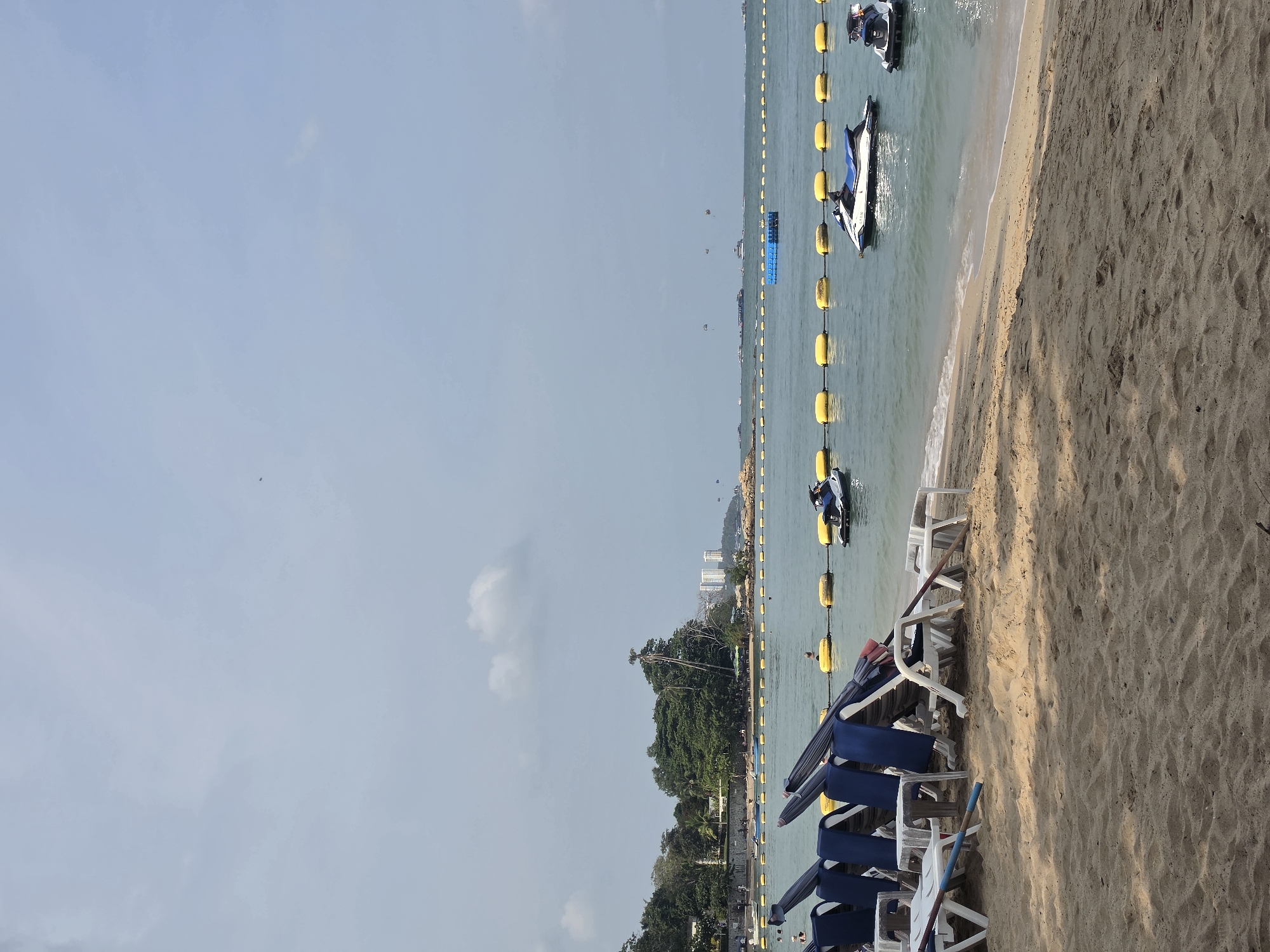 The image depicts a beach in Pattaya City, Thailand, with white sand in the foreground. Several rows of lounge chairs with blue umbrellas are arranged on the sand. The ocean is visible, delineated by a series of yellow buoys connected by rope, which act as a boundary line. Several jet skis are anchored in the water beyond the buoys. In the distance, a green, hilly landmass is visible, with buildings identifiable as part of the city skyline. The sky is mostly clear with a few scattered clouds.