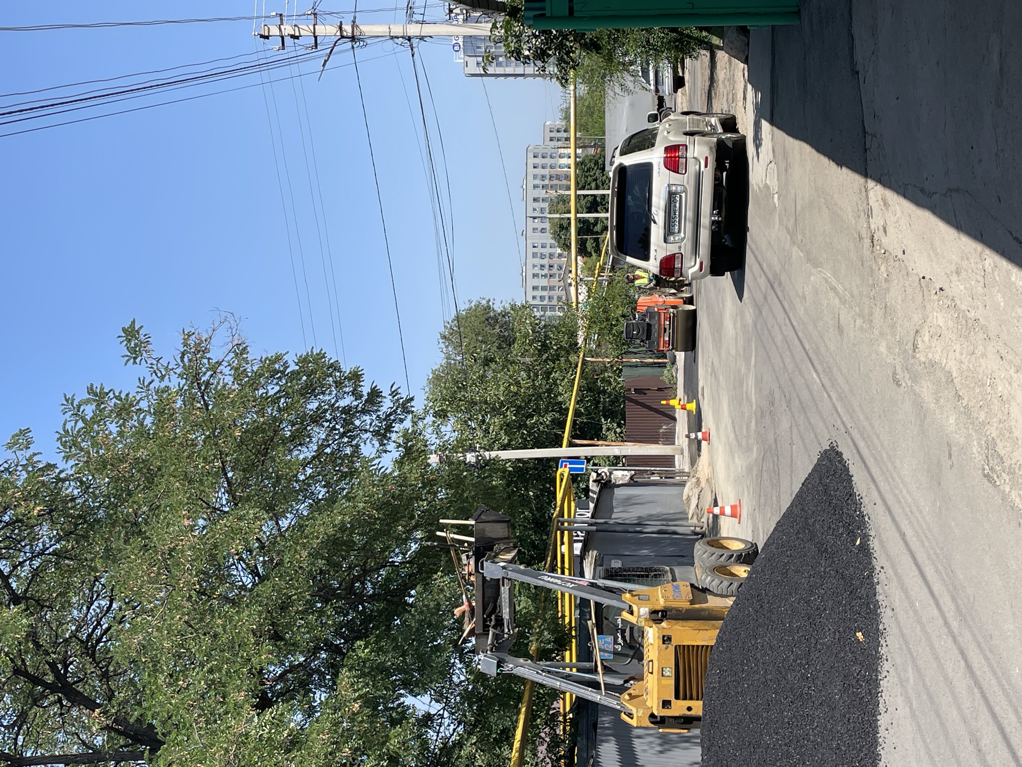 An asphalt paving project is underway on a street in Almaty, Kazakhstan. A pile of asphalt is visible on the left, next to a yellow forklift. A white SUV is parked on the right side of the road. In the background, a small orange roller is being operated by a worker wearing a reflective vest. Several residential buildings are visible in the distance. The sky is clear and blue.
