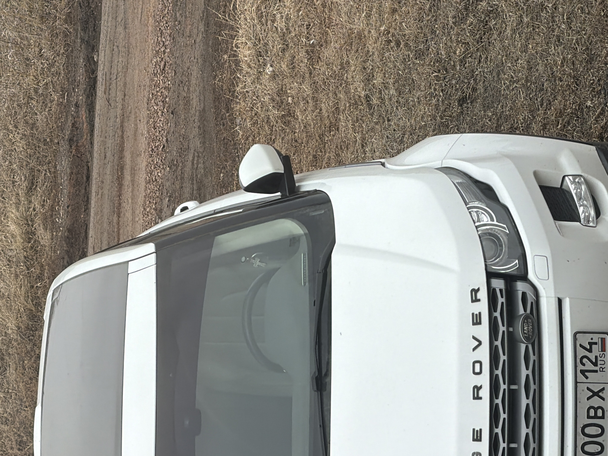 The image shows the front of a white Range Rover SUV parked on a dirt road next to dry grass. The vehicle has a Russian license plate reading "000BX 124 RUS," indicating the Krasnoyarsk region of Russia. No people are visible in the image.