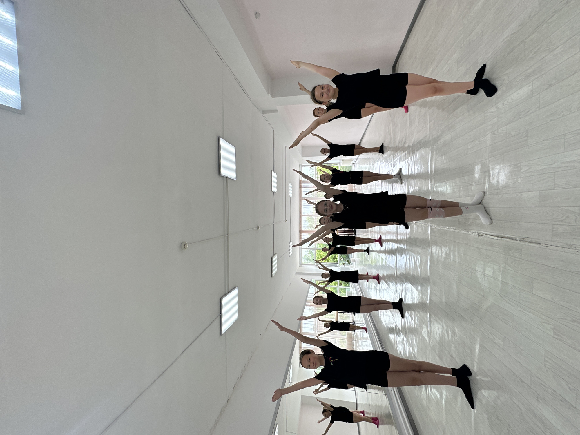The image depicts a group of young girls in a dance studio, likely in Votkinsk, Russia, practicing a synchronized movement. They are all wearing black leotards or t-shirts and shorts, and many are wearing pink or white socks. Each girl has her arms raised above her head, forming a cohesive line across the room. The studio appears to be a professional dance space, with a large mirror lining one wall and fluorescent lights on the ceiling. The floor is light-colored and polished, reflecting the dancers and the environment. The overall atmosphere suggests a focused and disciplined dance class.