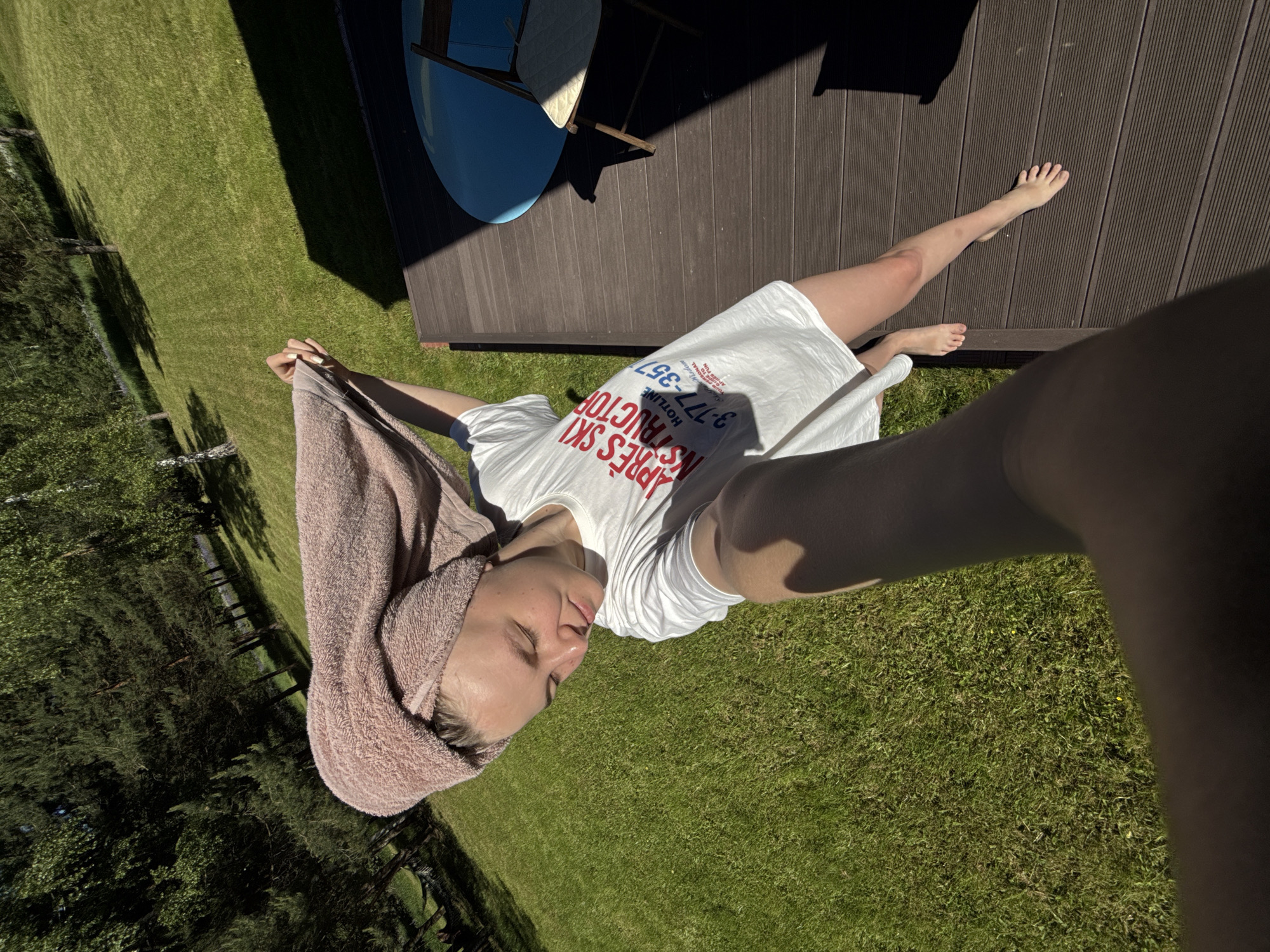 The image captures a young woman standing on a wooden deck outdoors in Копнино, Russia, under bright sunlight. She is taking a selfie, holding the camera in her outstretched right hand, which is prominently featured in the lower left foreground. Her head is wrapped in a light pink terry cloth towel. She is wearing a loose white t-shirt with red lettering spelling "APRES SKI INSTRUCTOR" and blue text that includes "HOTLINE 3-177-357". Her bare legs and feet are visible, with her left foot resting on the deck and her right foot slightly lifted. Her gaze is directed downwards, and her expression is tranquil.
The dark brown wooden deck occupies the right and lower sections of the frame. A deep, angular shadow from an unseen structure extends across the deck, obscuring a blue circular table and a wooden chair with a light cushion positioned in the upper right. Beyond the deck, a lush, green grassy lawn slopes gently upwards into the background. Several trees, including light-barked birches and darker conifers, are scattered across the distant landscape, with shadows cast upon the grass. The scene is brightly illuminated, with clear distinctions between sunlit areas and sharp, dark shadows.