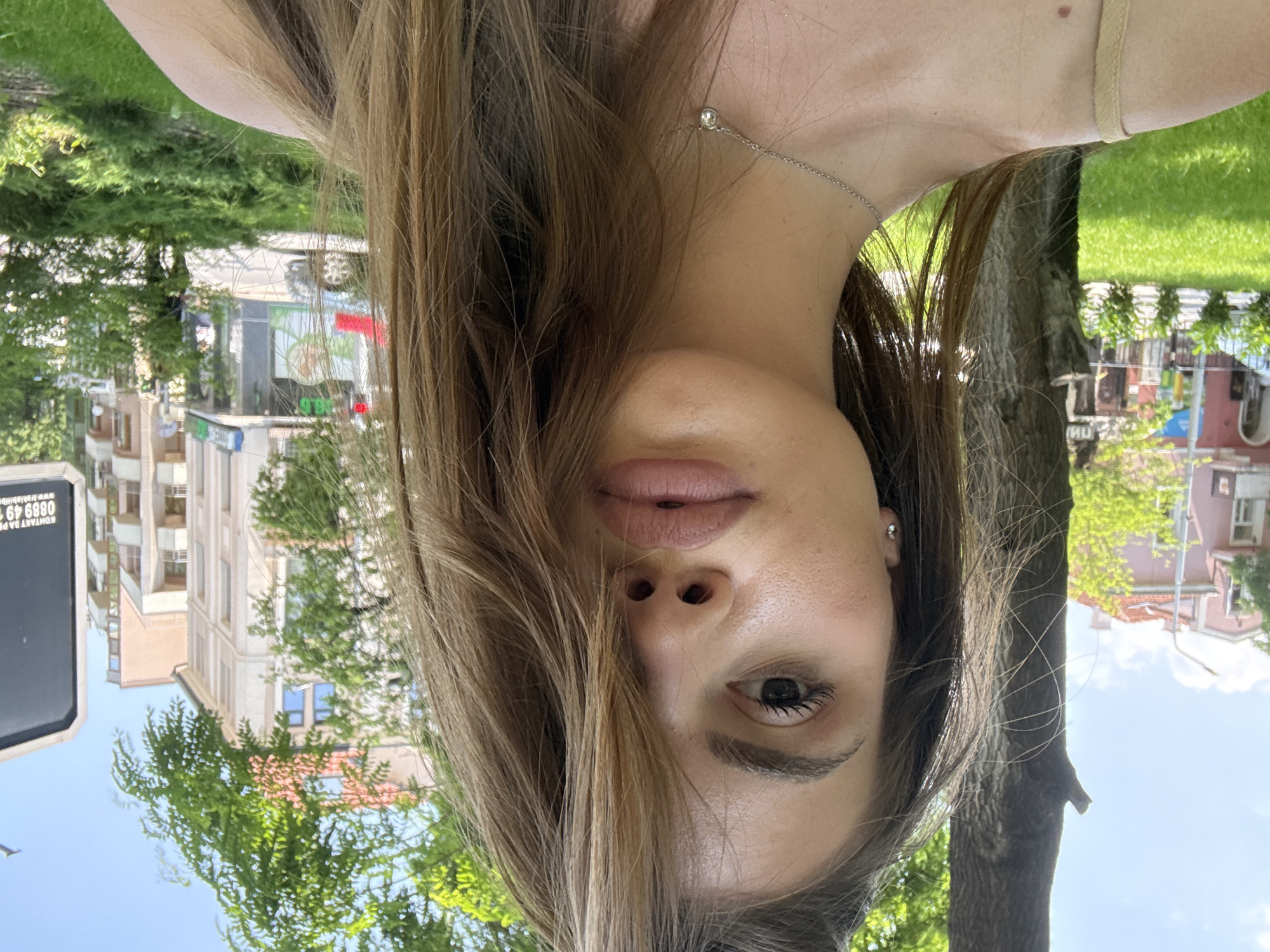 The image is an upside-down, close-up selfie of a young woman, set outdoors in Plovdiv, Bulgaria, on a bright, sunny day. The woman is centered in the frame, looking directly at the camera with a neutral expression. Her long, light brown hair, with some lighter strands, cascades around her face, partially obscuring her forehead and mouth. She wears subtle eye makeup, including mascara and defined eyebrows, and natural-toned lips. A delicate silver necklace with a small, clear stone pendant adorns her neck, and a small silver stud earring is visible in her left ear. A light-colored strap, possibly from her clothing, is visible on her right shoulder.
The background is a vibrant, out-of-focus urban park or street scene, featuring lush green trees, grass, and buildings under a clear blue sky. Residential or commercial buildings with windows and balconies are visible on the left side of the frame. A prominent tree trunk stands to the right of the woman.
Notable visible text includes a partially obscured white rectangular sign displaying "KOHTAKT" (Kontakt), a phone number "0889 49 6880," and a website "www.trafikbid.eu." Another green sign on a building in the background shows "100" and "OTB." The scene conveys a relaxed, outdoor moment captured in a vibrant urban environment.