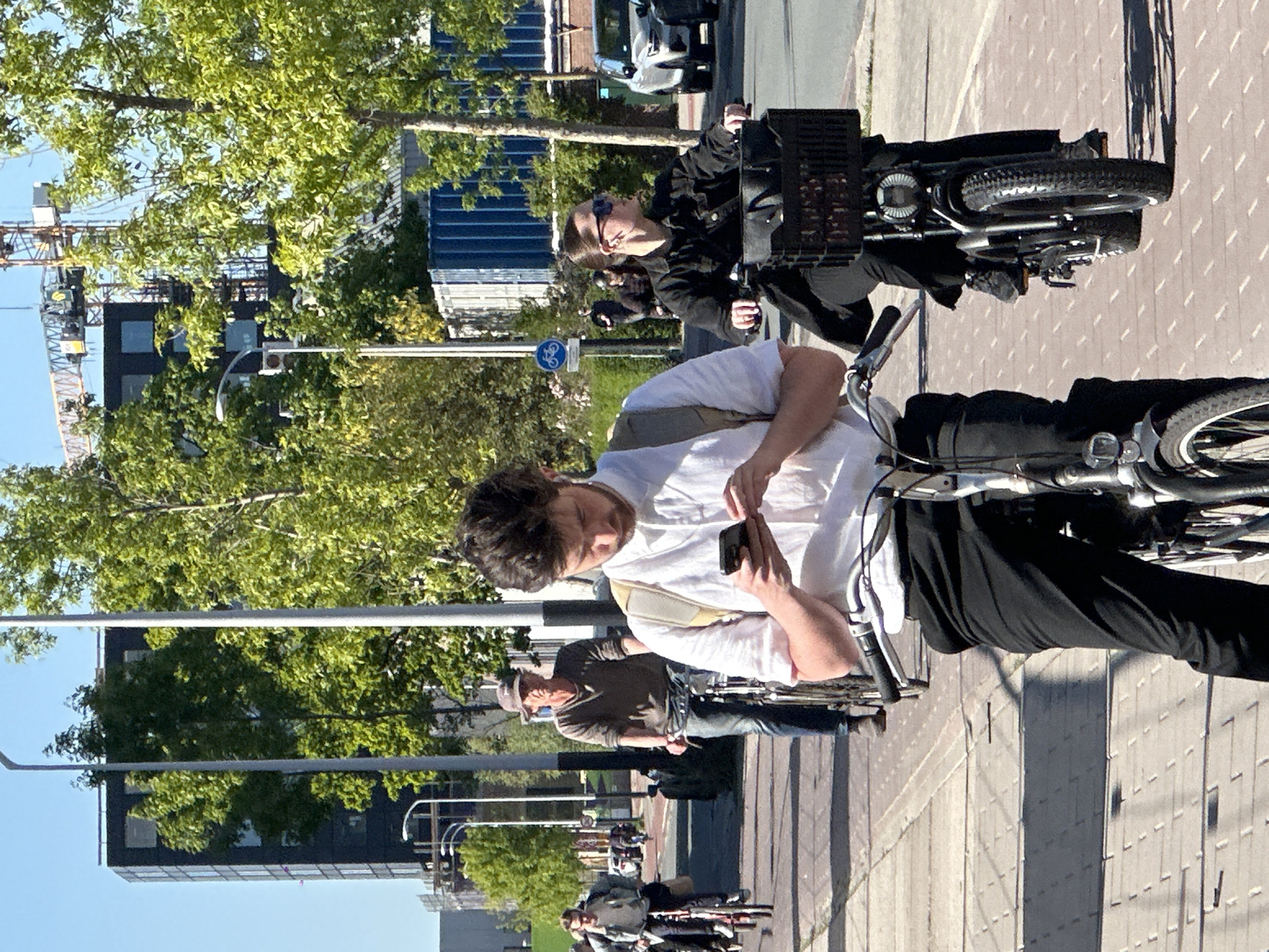 This image captures a vibrant urban street scene, likely in Amsterdam, bathed in bright sunlight on a clear day. The setting features multiple cyclists, modern buildings, and green trees.
In the foreground, a person with dark hair, wearing a white shirt and dark pants, is seated on a bicycle. They are actively engaged with a smartphone held in their hands, looking down at the screen. A light-colored shoulder bag is visible across their body. Behind them, another individual wearing a black jacket and sunglasses is also on a bicycle, possibly an e-bike, which has a black storage crate mounted on its front. Further back, additional cyclists, including one wearing a dark top and a cap, are visible.
The scene suggests a pause in cycling activity, perhaps at a junction or a waiting spot. In the background, modern buildings with dark exteriors and large windows are partially visible through the foliage of trees. To the left, a tall construction crane stands against the clear blue sky, indicating ongoing development. The ground is paved with light-colored blocks or tiles. A blue circular sign featuring a white bicycle symbol is discernible, denoting a mandatory cycle path, a common sight in Dutch cities. The strong sunlight casts crisp shadows, suggesting the time of day is midday or early afternoon. No alphanumeric text is clearly legible within the image, apart from the symbolic bicycle sign.