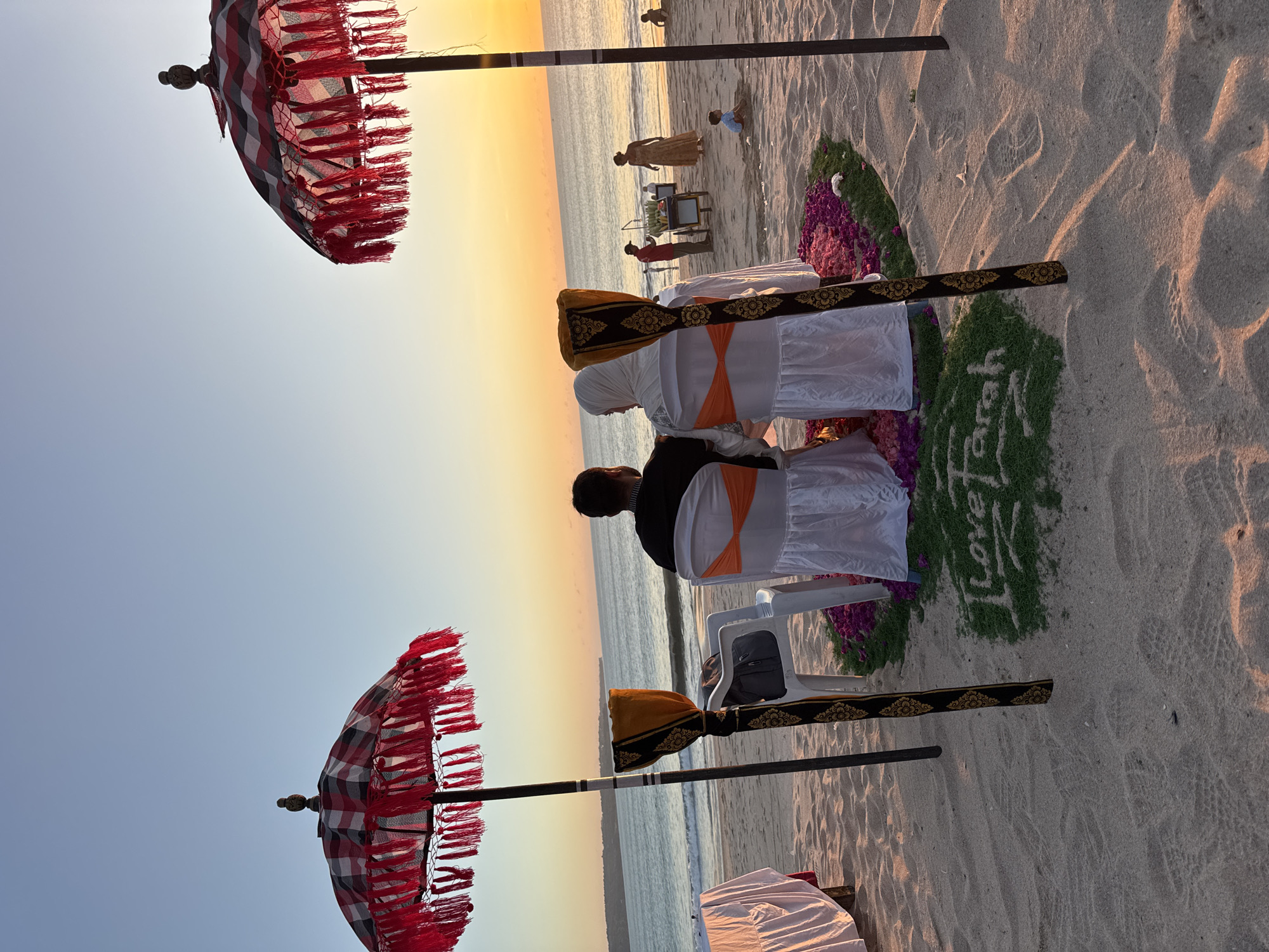 The image depicts a serene beach ceremony in Kuta, Indonesia, at either sunrise or sunset, characterized by the warm orange and yellow glow on the horizon and the clear, light blue sky above. The sandy beach extends into the mid-ground, meeting the calm ocean with gentle waves.
In the foreground and mid-ground, the primary subjects are two individuals seated on white chairs with orange sashes, facing the ocean with their backs to the viewer. One person wears a black top, while the other is dressed in white and wears a light-colored headscarf. Two traditional Balinese ceremonial umbrellas (payung tedung) with red fringes and a black, white, and red plaid pattern flank the seated couple. Two additional unoccupied white chairs, similarly decorated with orange sashes and draped with a black and gold patterned fabric, are positioned closer to the camera.
Between the seated couple and the water, a decorative arrangement of green foliage and purple flowers is laid out on the sand. Within this arrangement, the words "love Farah" are clearly spelled out in white. In the background, closer to the waterline, several other people are visible, including a person standing near a small cart and two smaller figures on the sand. The setting suggests an intimate and romantic event.