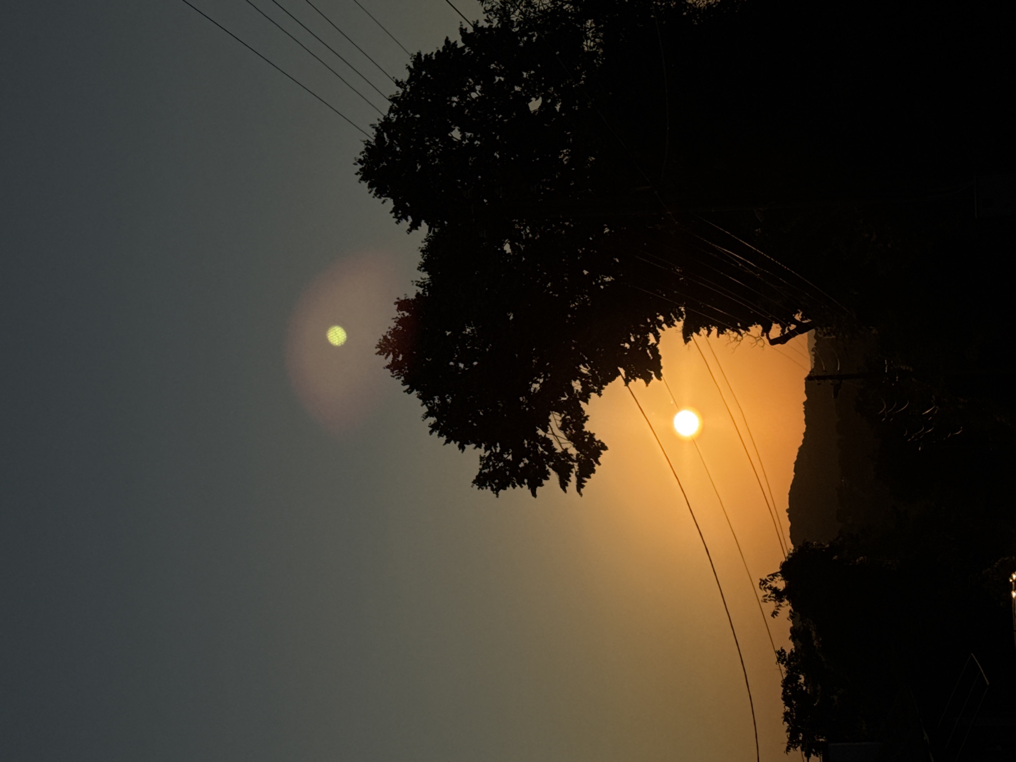 The image captures a dusky sky over Krushevo, Bulgaria, with the sun setting behind silhouetted hills and trees. Two distinct orbs of light are visible in the sky; the lower, larger orb is a bright orange sun, while the upper, smaller orb is a pale yellow, possibly a lens flare or another celestial body. Power lines stretch across the frame, casting faint diagonal lines against the darkening sky. A large tree, silhouetted in black, dominates the right side of the image, its branches reaching upwards. The foreground is a dark, indistinct landscape, hinting at urban structures or vegetation. The overall mood is tranquil and atmospheric, characteristic of twilight.