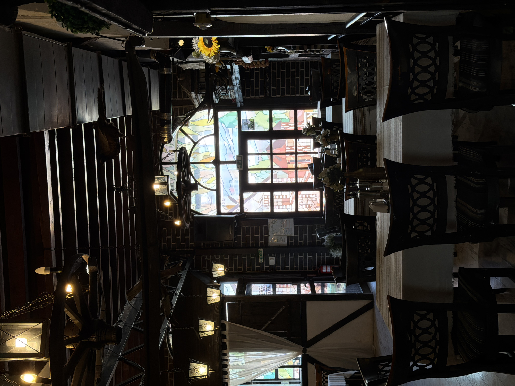 The image depicts the interior of an unoccupied traditional-style restaurant or tavern, located in the city of Varna, Bulgaria. The setting is characterized by a rustic aesthetic, featuring exposed dark wooden ceiling beams and dark brick walls. Numerous dark wooden chairs with elaborately patterned, possibly carved, backrests are arranged around rectangular tables, each covered with a white tablecloth and a patterned fabric runner. Each table holds a small decorative vase containing artificial flowers or dried arrangements, alongside condiment sets.
Overhead, multiple chandeliers hang, including large wagon-wheel designs incorporating lantern-style lights and several smaller ornate lanterns. Illumination within the room is dim, supplemented by bright natural light filtering through two large windows on the far wall. The most prominent window is an arched structure featuring a vibrant stained-glass depiction of a colorful townscape. Below this, rectangular windows also offer glimpses of the bright exterior, obscured by the stained glass art. To the left, another window is partially covered by a tied-back sheer white curtain, revealing an exterior view.
Additional decor includes a mounted set of deer antlers near the main arched window and a string of dried red peppers or garlic hanging on the right wall. An artificial sunflower in a vase and a small artificial green plant are visible on shelves to the right. A green "EXIT" sign is mounted above a dark wooden doorway on the left wall, next to which a red fire extinguisher is visible on the floor. A white informational sign is also affixed to the brick wall near the exit. No individuals are present in the scene.