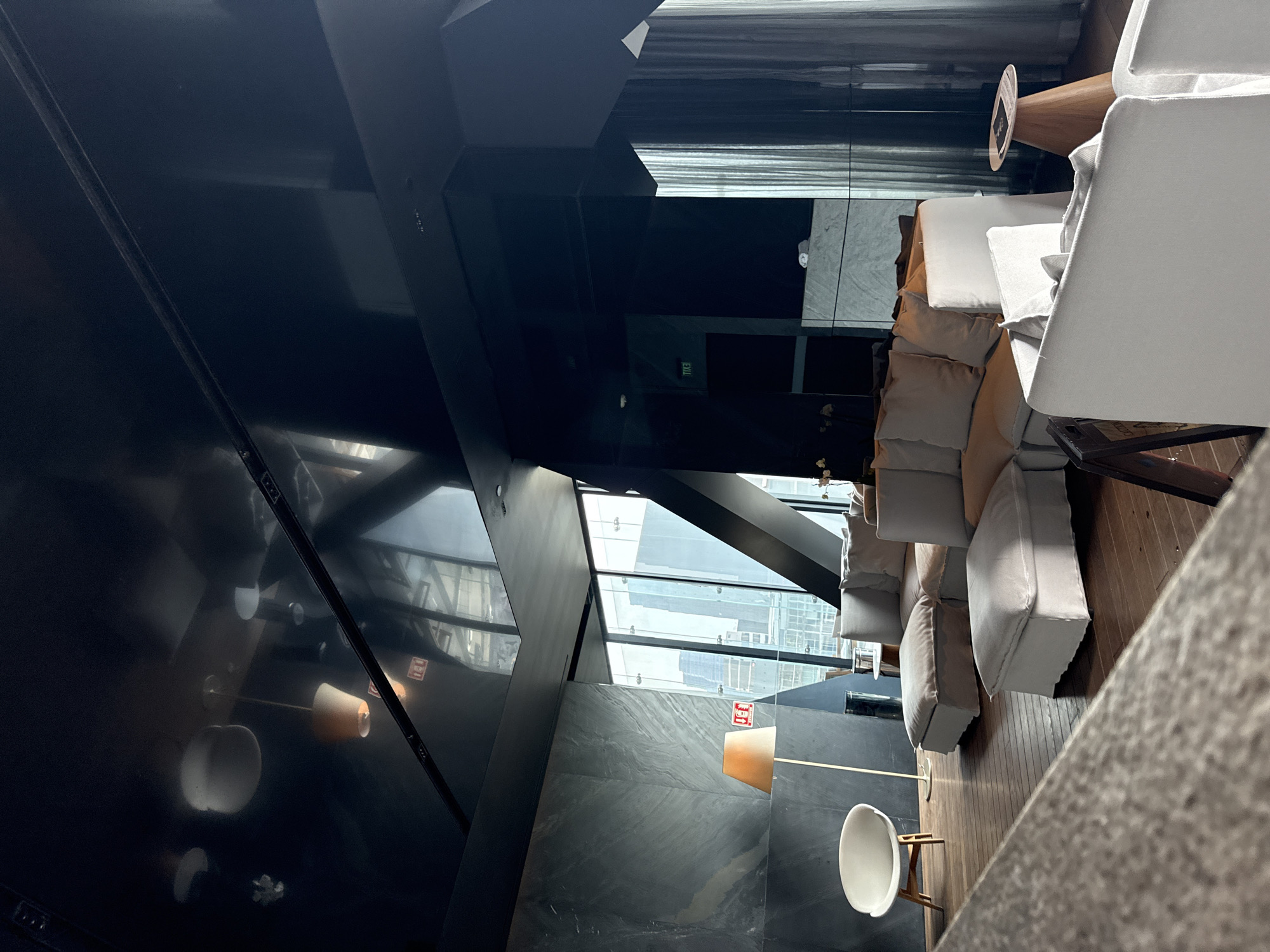 The image shows an interior view of a building located in Mexico City, Mexico, during daytime.
The building features modern architectural design with a striking geometric ceiling and large glass windows providing a view of the cityscape. The interior appears to be a lounge or waiting area, furnished with a white chair, a long sofa with light-colored cushions, and a wooden side table.
The floors are wooden, and the lighting is provided by floor lamps and possibly recessed ceiling lights. The building’s structure and glass panels allow ample natural light to enter the space. The scene is quiet and empty of people, suggesting a moment of inactivity in the space.