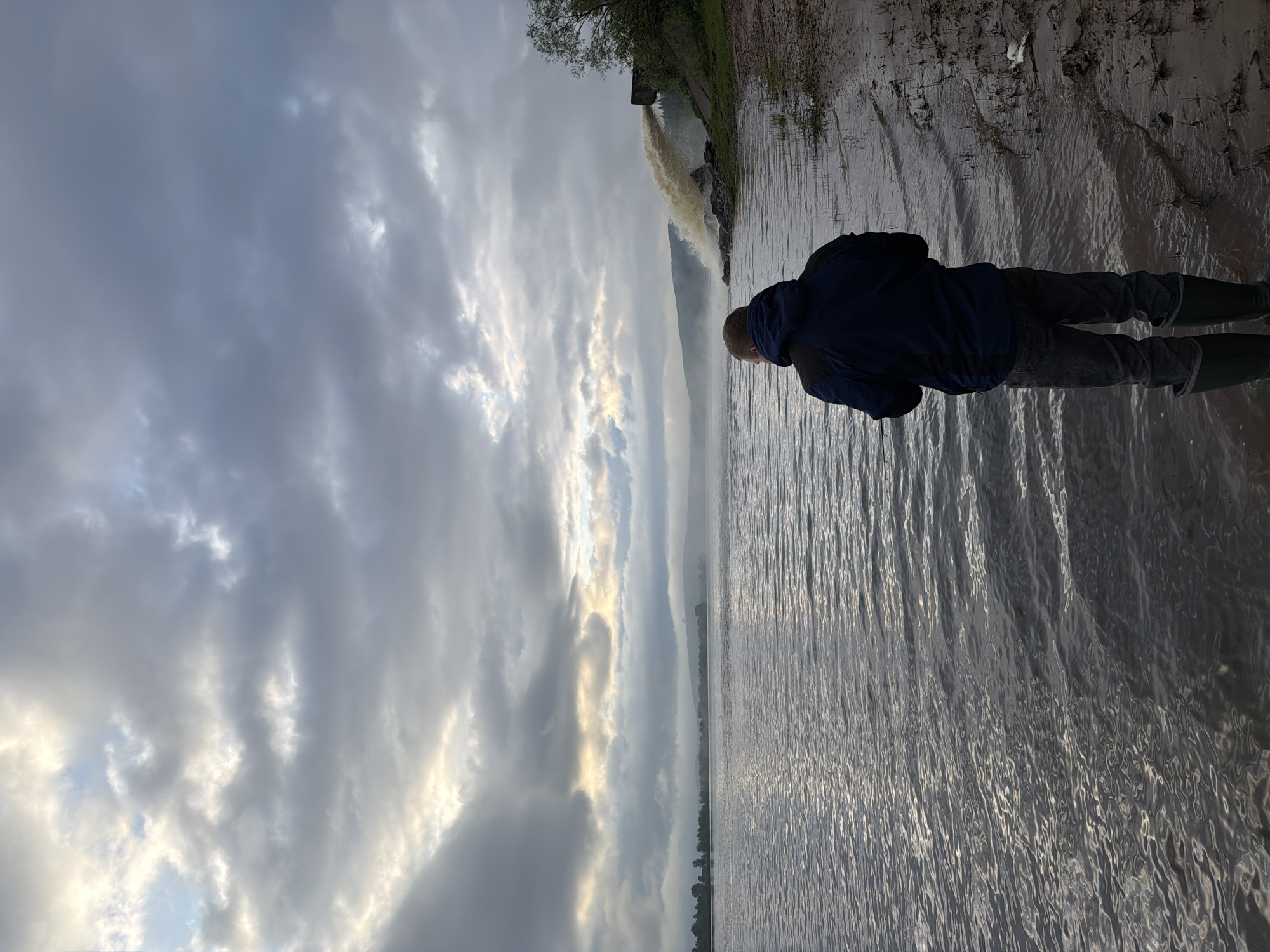 A person stands at the edge of a body of water under a cloudy sky. The person is wearing a dark blue jacket, dark pants, and tall boots. The water is dark and rippled. To the right of the person, the shoreline is muddy with sparse vegetation. In the distance, water is being discharged from a structure. The location is likely Ol'khovskoye, Russia.