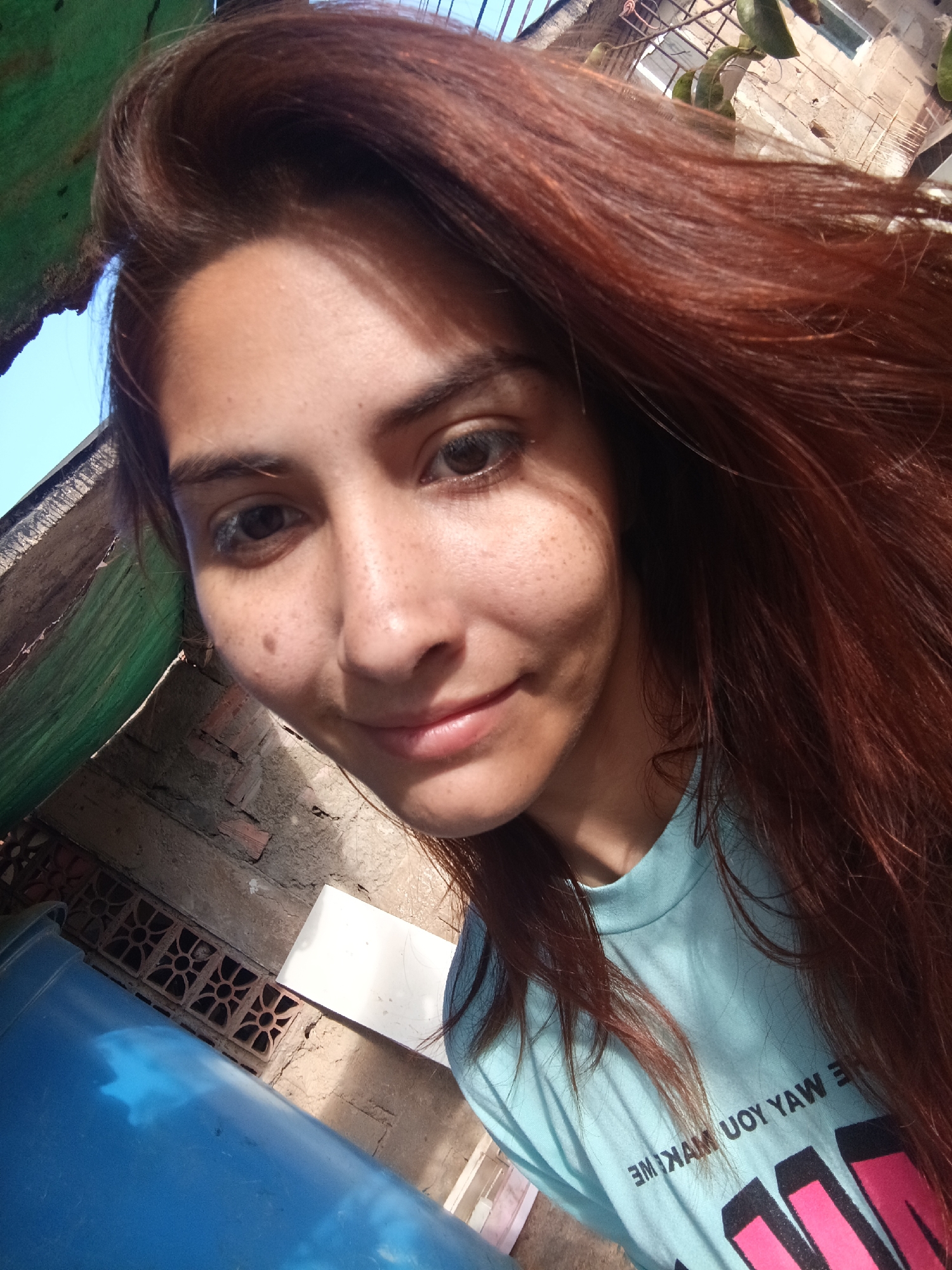 A selfie captures a woman with auburn hair and brown eyes, smiling gently at the camera. Her skin exhibits freckles, and she wears a light blue t-shirt featuring reversed text. The backdrop reveals an exterior setting characterized by weathered brickwork, a corrugated green structure overhead, a blue plastic container, and a glimpse of the sky. Due to the limited field of view, pinpointing the exact location within Juan Griego, Venezuela, based solely on the visual elements, is not possible.