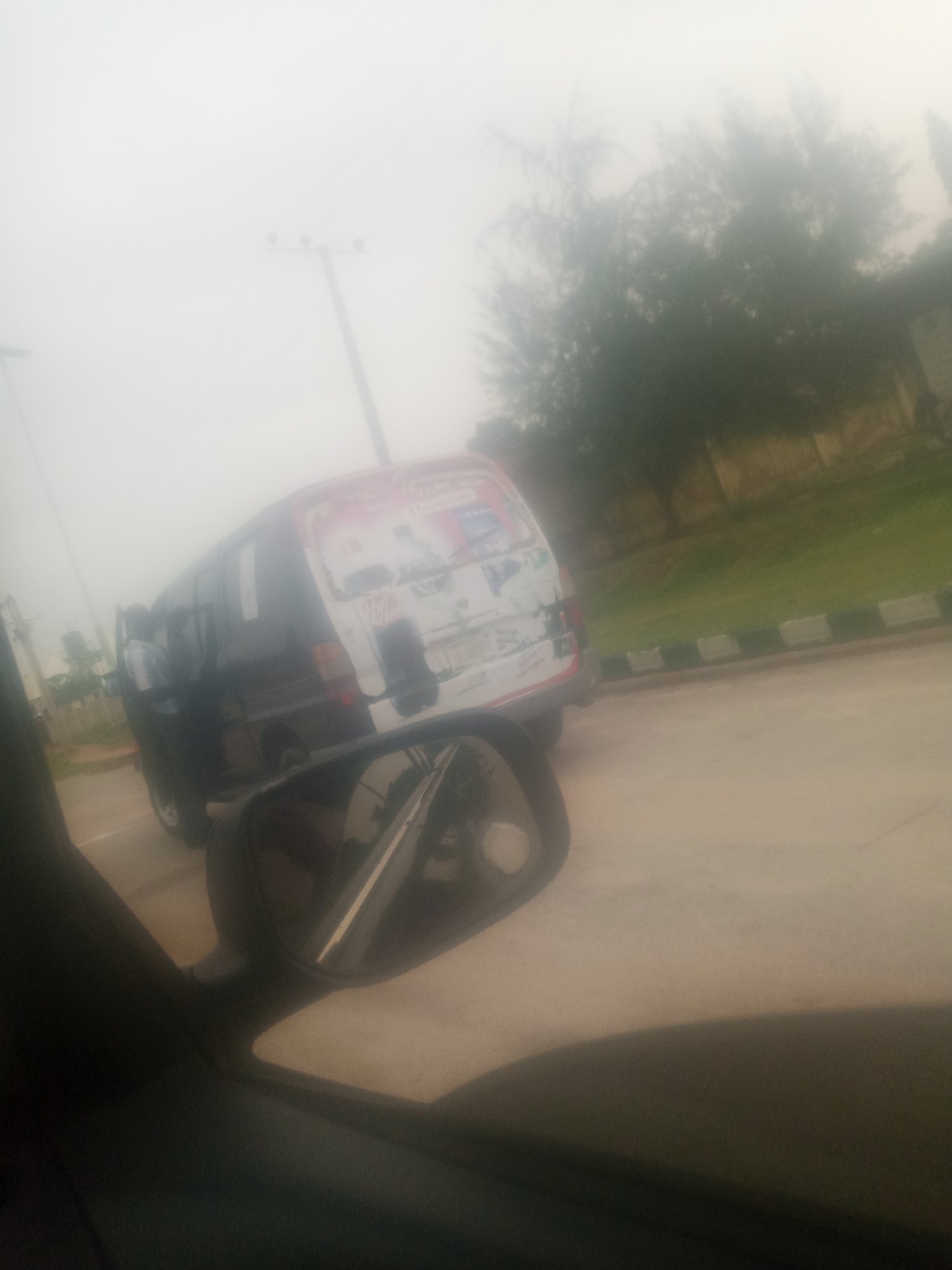 An overcast day in Jalingo, Nigeria. A dark-colored van, possibly a passenger bus, is parked on the right side of the road. The van has some graphics and text on its rear, though they are indistinct due to the lighting and focus. A side-view mirror of another vehicle is visible in the foreground, reflecting a portion of the van. The road appears to be paved, with a grassy area and a low barrier to the right of the van. Trees and a utility pole are visible in the background, partially obscured by fog or haze. The scene suggests a moment captured from inside a vehicle.