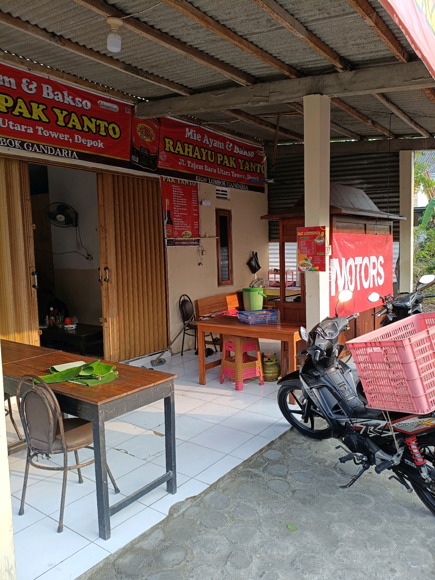 This image depicts an outdoor scene in Maguwoharjo, Indonesia, likely a food stall or small restaurant. The foreground shows a rustic wooden table set with banana leaves, next to a dark metal chair. To the right, a black motorcycle is parked, equipped with a large pink basket. Further back, there's a wooden stall structure with a red banner displaying the word "MOTORS" in white text. Several tables and chairs are visible, suggesting a seating area. The background includes a building with corrugated metal roofing supported by wooden beams and concrete pillars. Signs for "PAK YANTO" and "Mie Ayam & Bakso" are prominent, indicating the establishment's offerings. The overall environment appears to be a casual, open-air dining space.